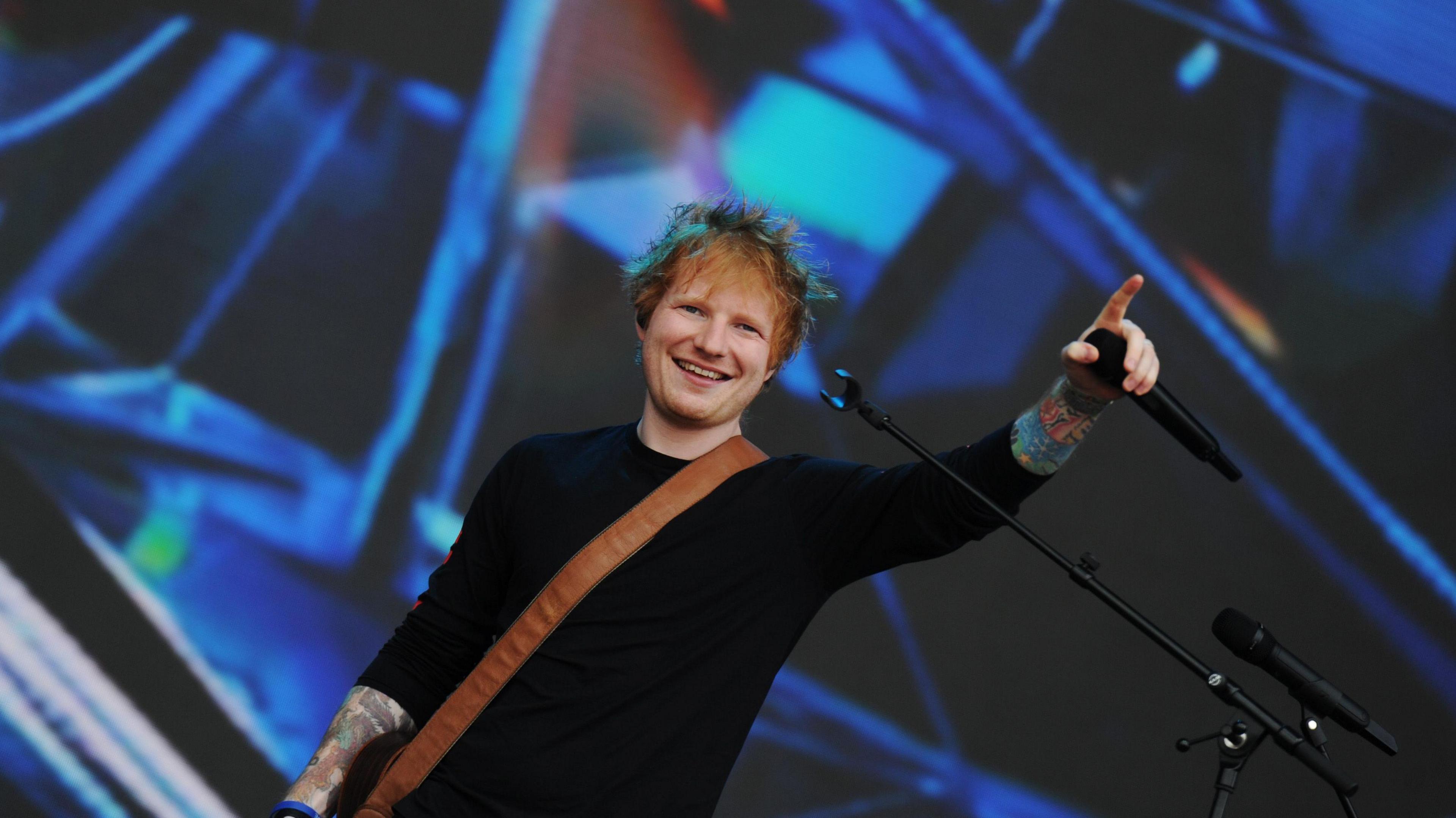 Ed Sheeran is pictured on stage at the Radio 1 Big Weekend festival in 2022. He is smiling while holding a black microphone in the air. He is wearing a black jumper and there is a brown leather guitar strap across his front. Blue and green shapes on the backdrop.