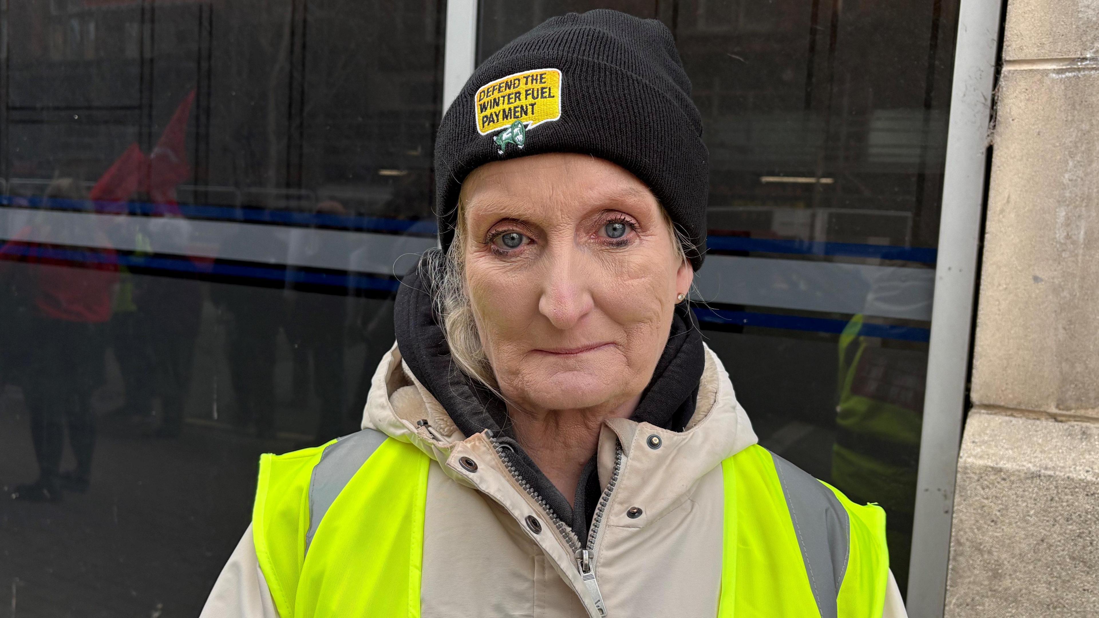 Rosalind is looking into the camera. She's wearing a high viz yellow jacket ad wearing a black beanie hat that says defend the winter fuel payment.