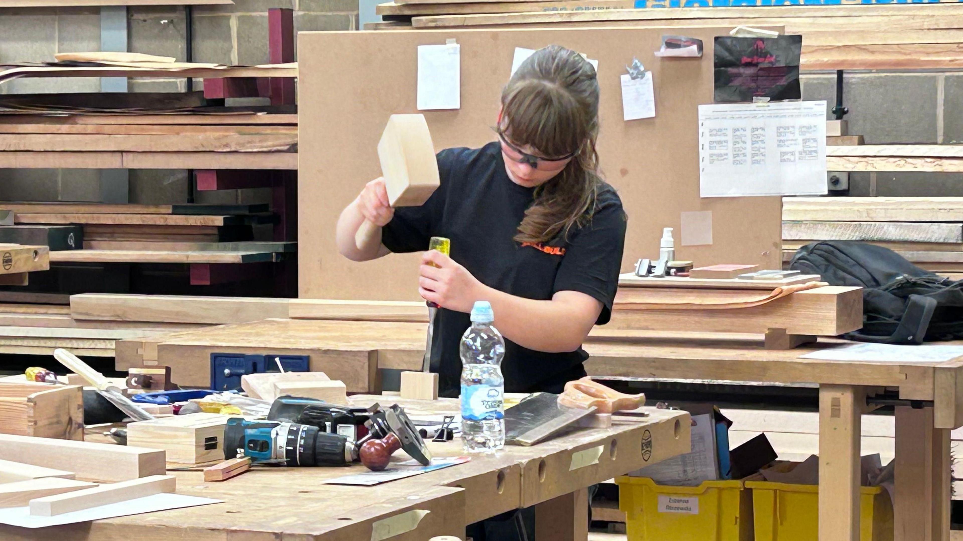 Maizie with long brown hair tied at the neck. She is wearing glasses - possibly safety goggles. She holds a wooden mallet in her right hand and a chisel in her left, and is working on a small piece of wood. There are other bits of wood and tools on a wooden bench in front of her. along with a bottle of water.