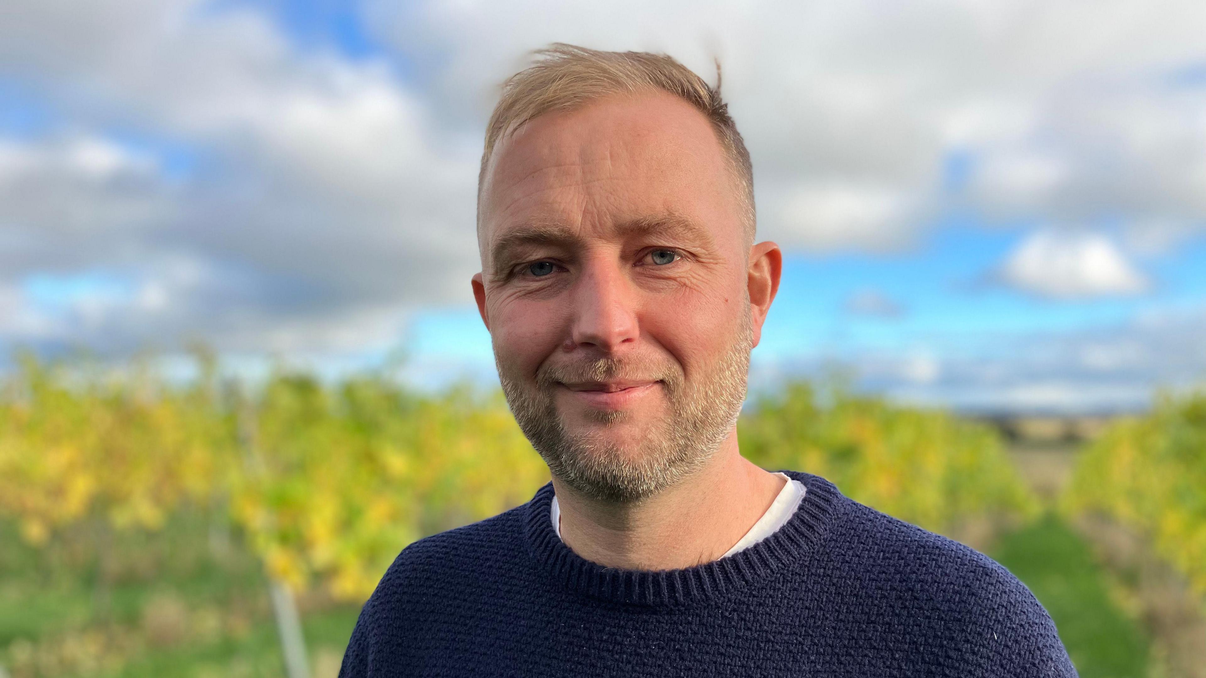 Man with thin blonde hair and a stubly beard is smiling while standing in a field of grapes. He is wearing a blue knitted shirt with a white shirt underneath