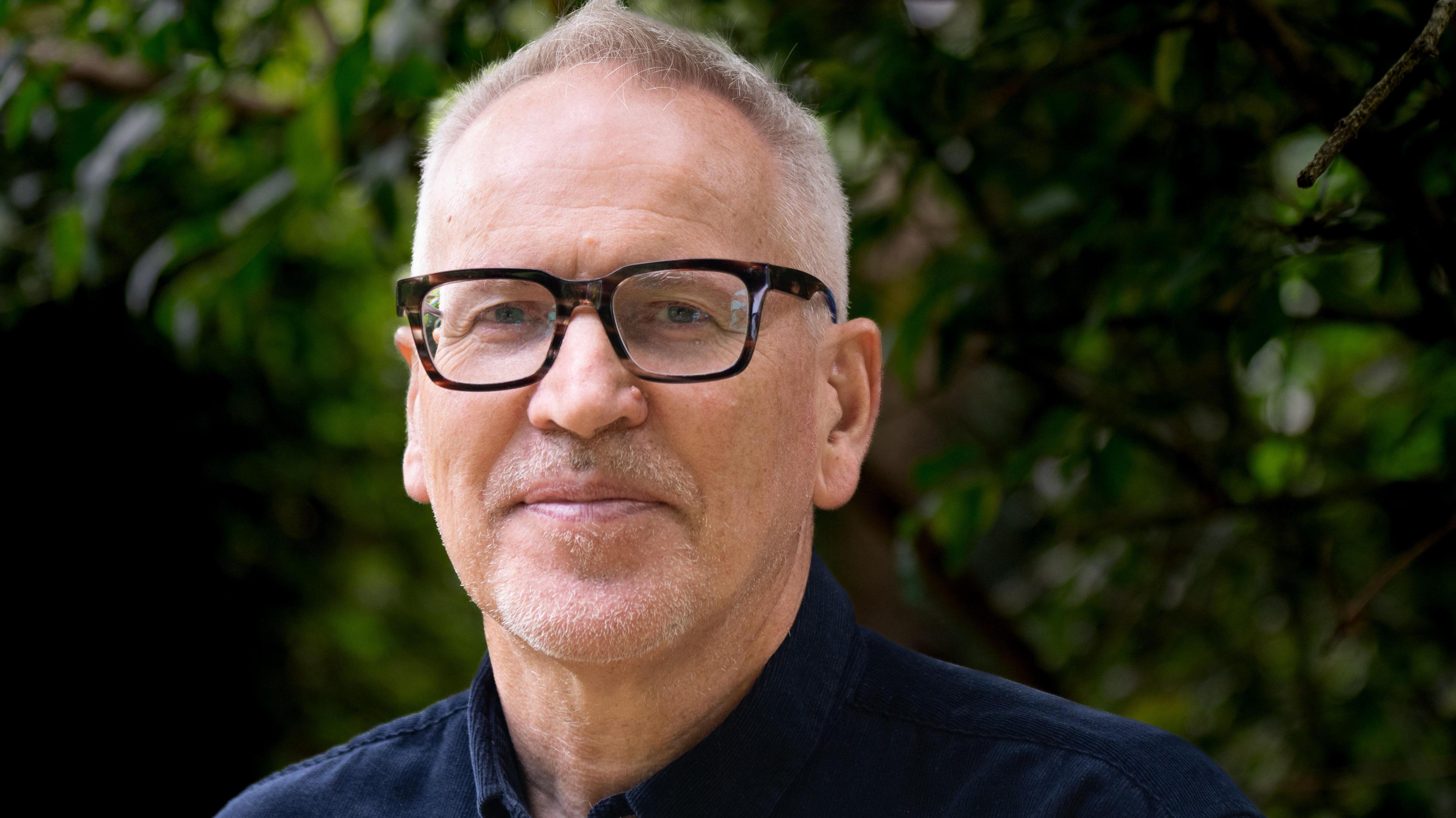 Ray Tostevin. He has short, light grey hair and stubble on his chin and upper lip. He is wearing glasses with a thick tortoiseshell frame. He is looking directly at the camera and smiling.