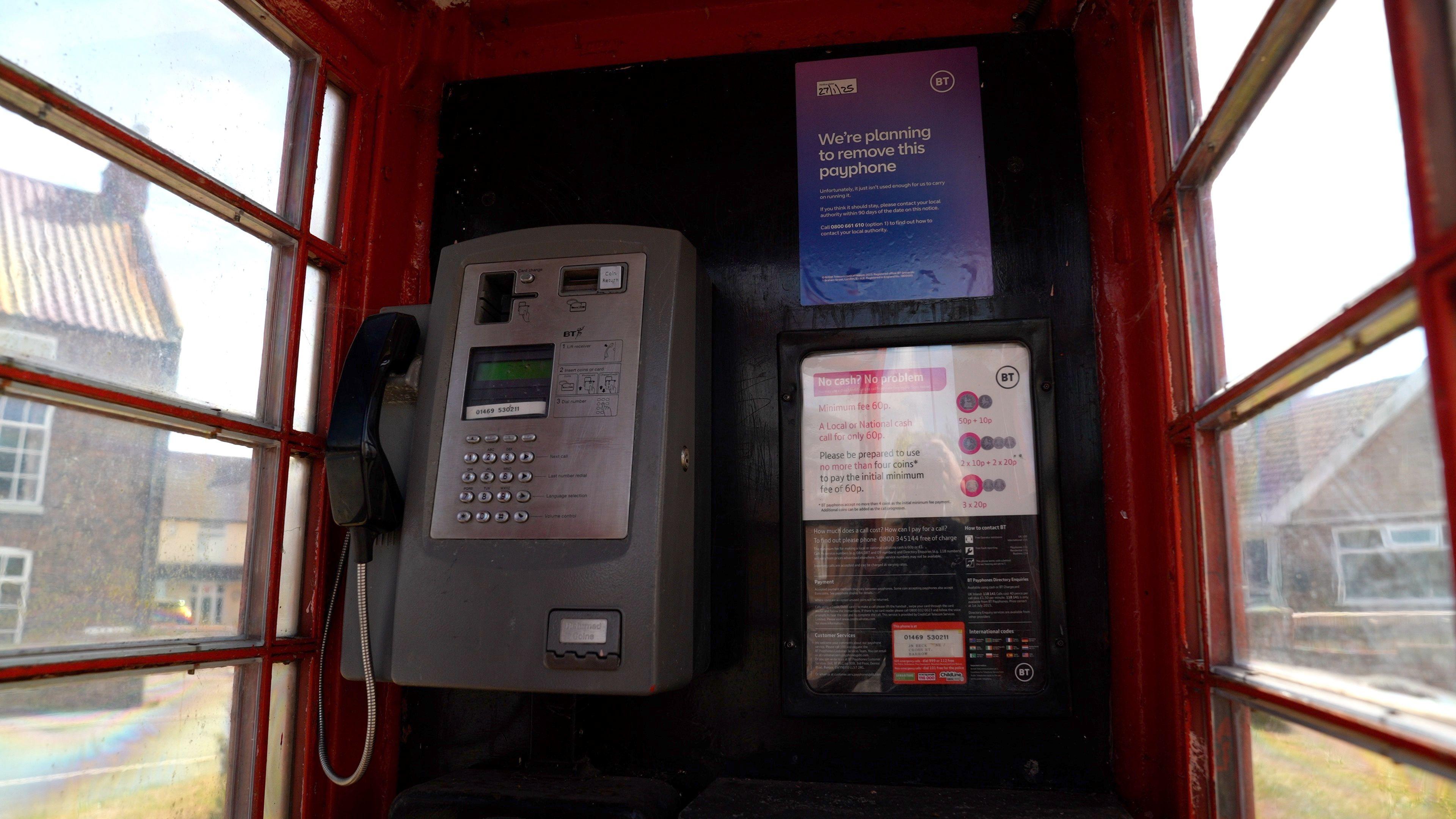 The inside of the Barrow upon Humber phone box with a grey payphone on the left side of the interior wall. There is a purple BT posted in the top right corner of the same wall which says 'we're planning to remove this payphone.' Below that is another BT poster, which is white and black. The phone box has see-through windows on either side and a red frame.