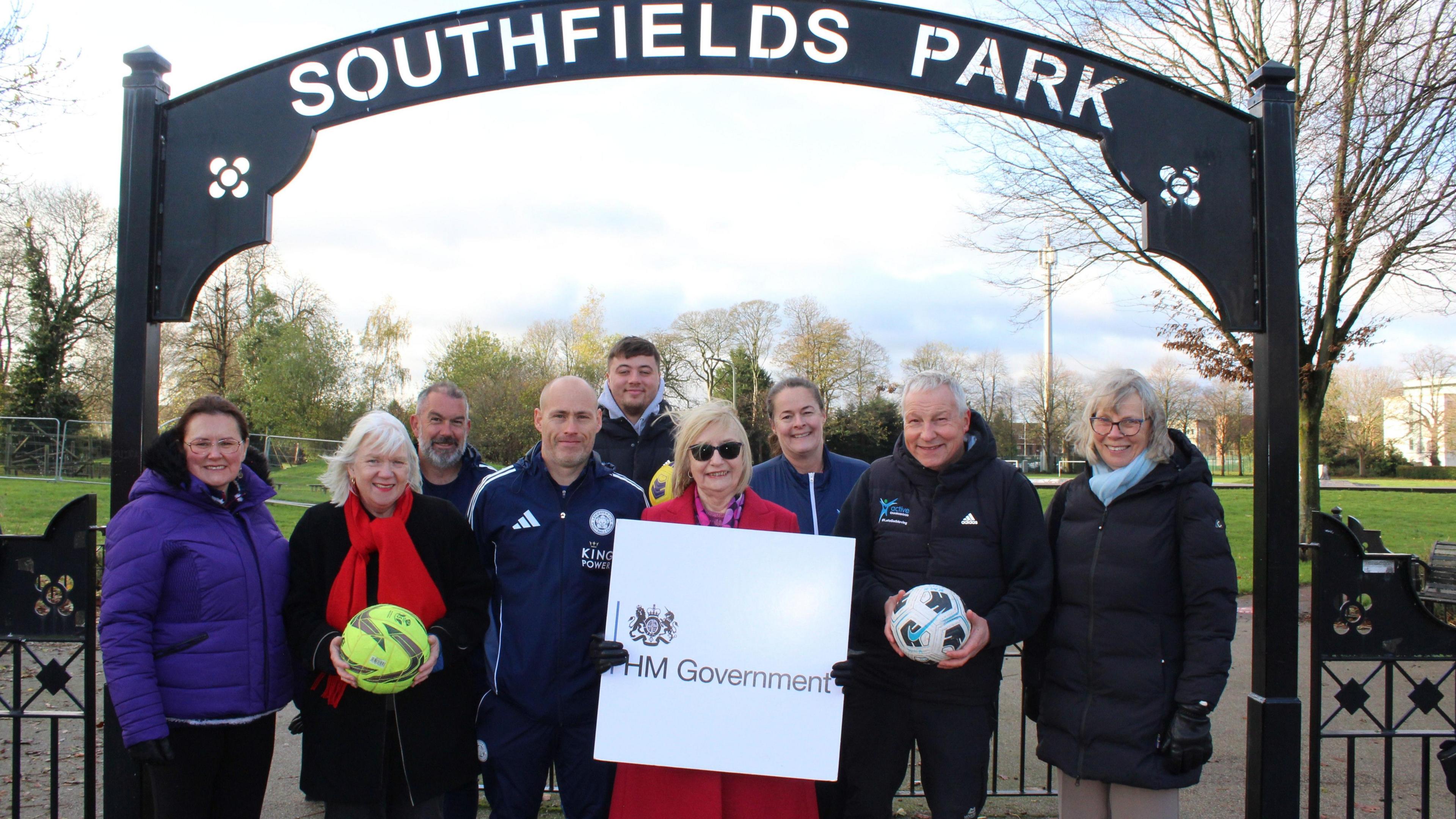 A group of people stood in front of a black iron arch which says Southfields Park on