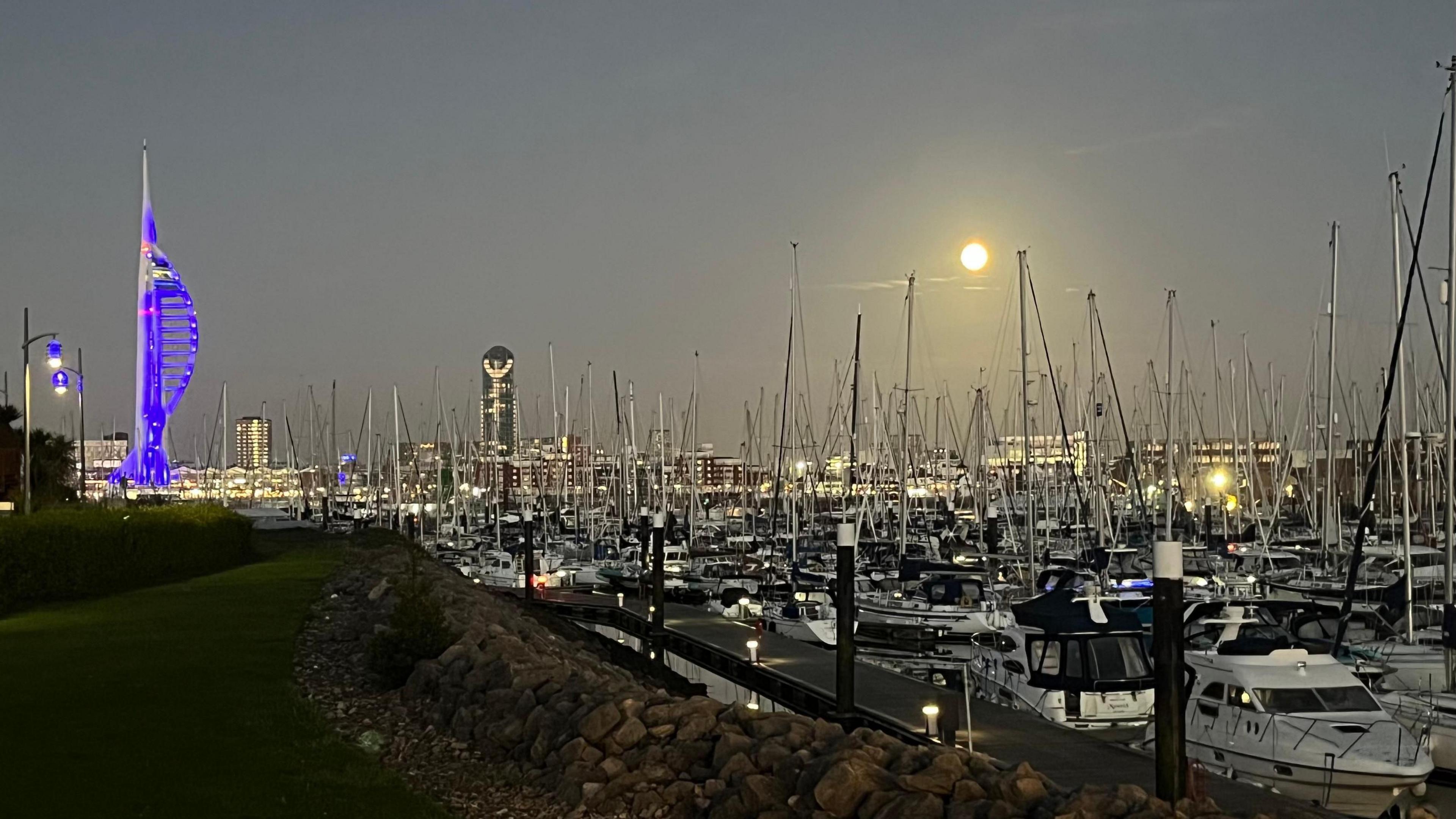 The Spinnaker Tower to the left of boats and the moon above the boats in the distant sky