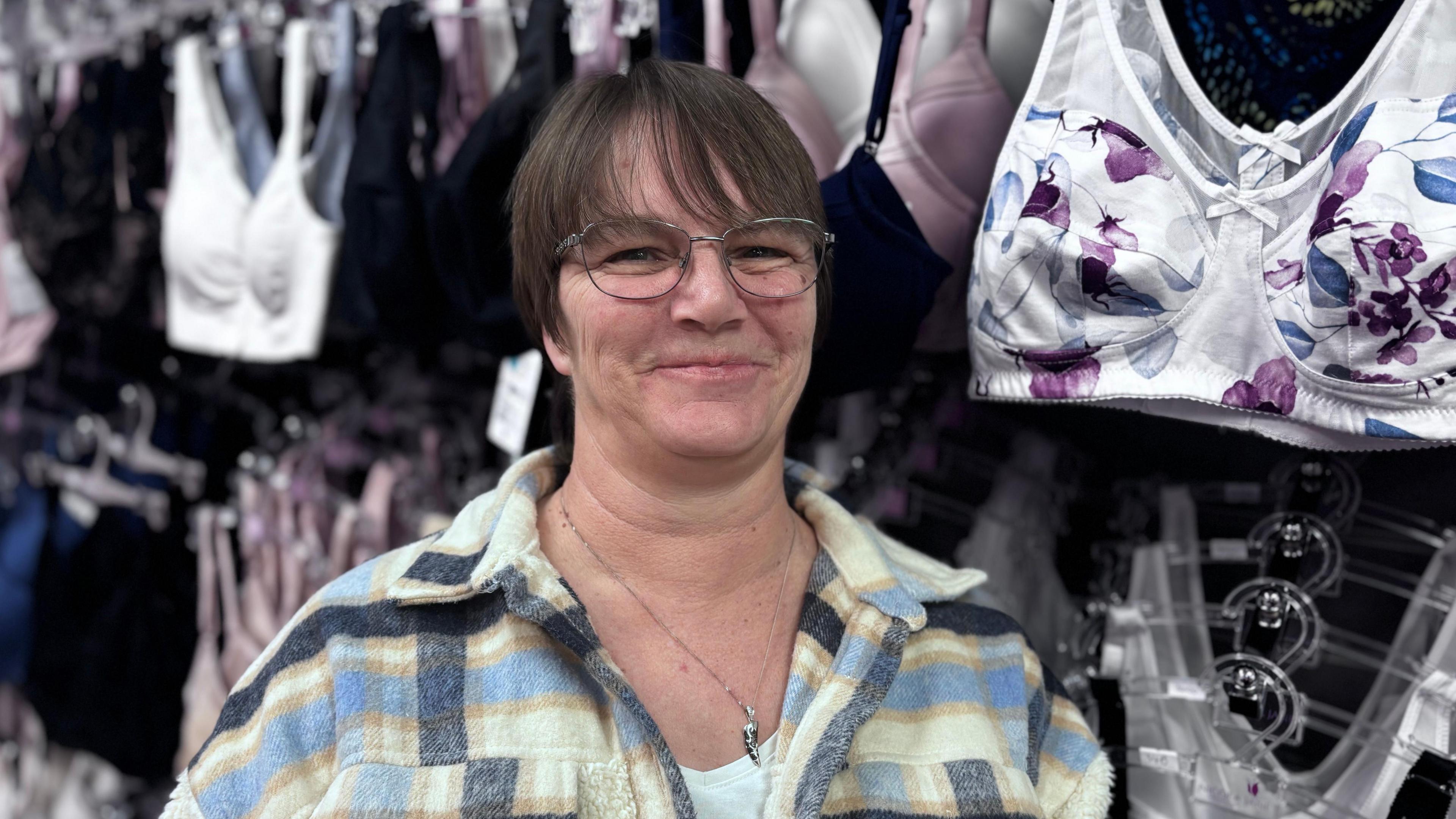 Woman with short brown hair and glasses, wearing a checked shirt, smiling at the camera in a lingerie shop. 