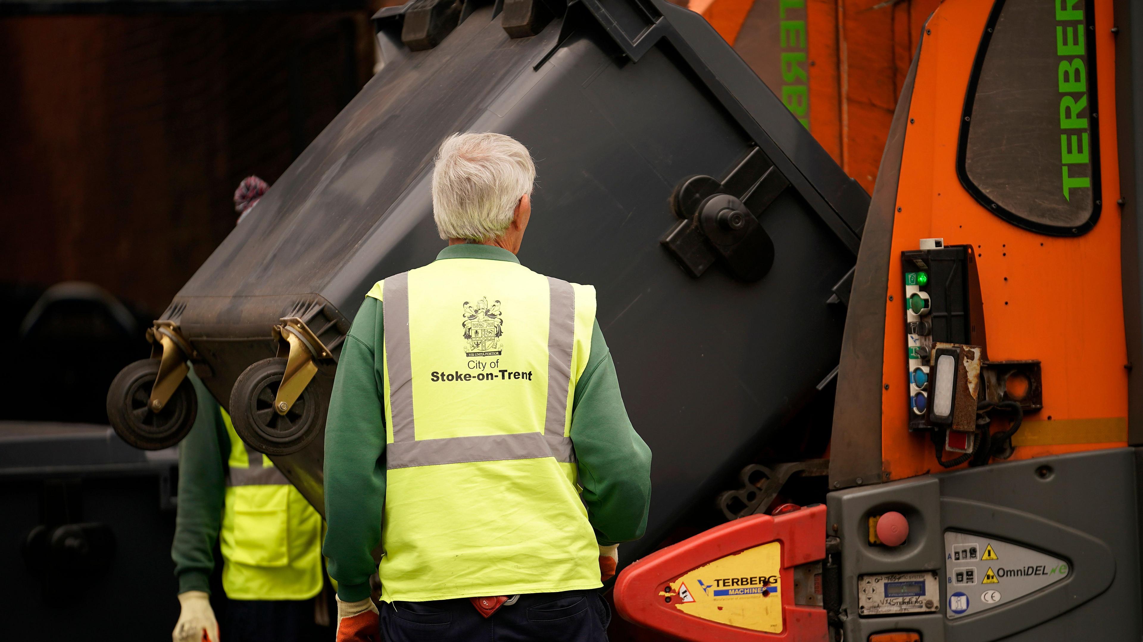 A bin lorry collects rubbish, flanked by two collectors wearing high-vis vests, in Stoke-on-Trent