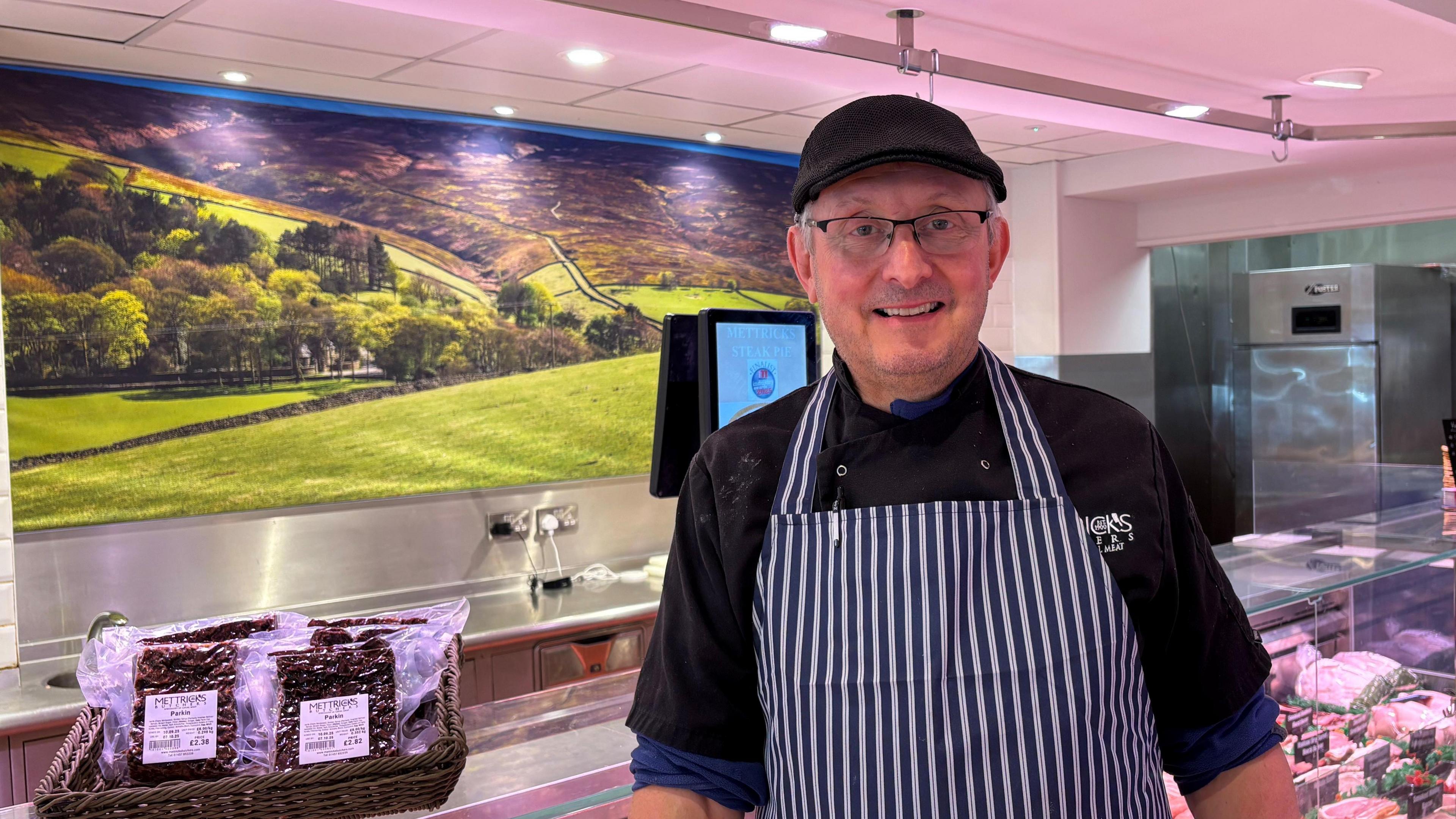 John Mettrick has a black flat cap and a blue and white striped butchers' apron on. He is wearing glasses and some grey hair is visible on the side of his head, under his cap. Behind him is a butchers' shop, with meats in cabinets and vacuum-packed goods in a basket on the counter.