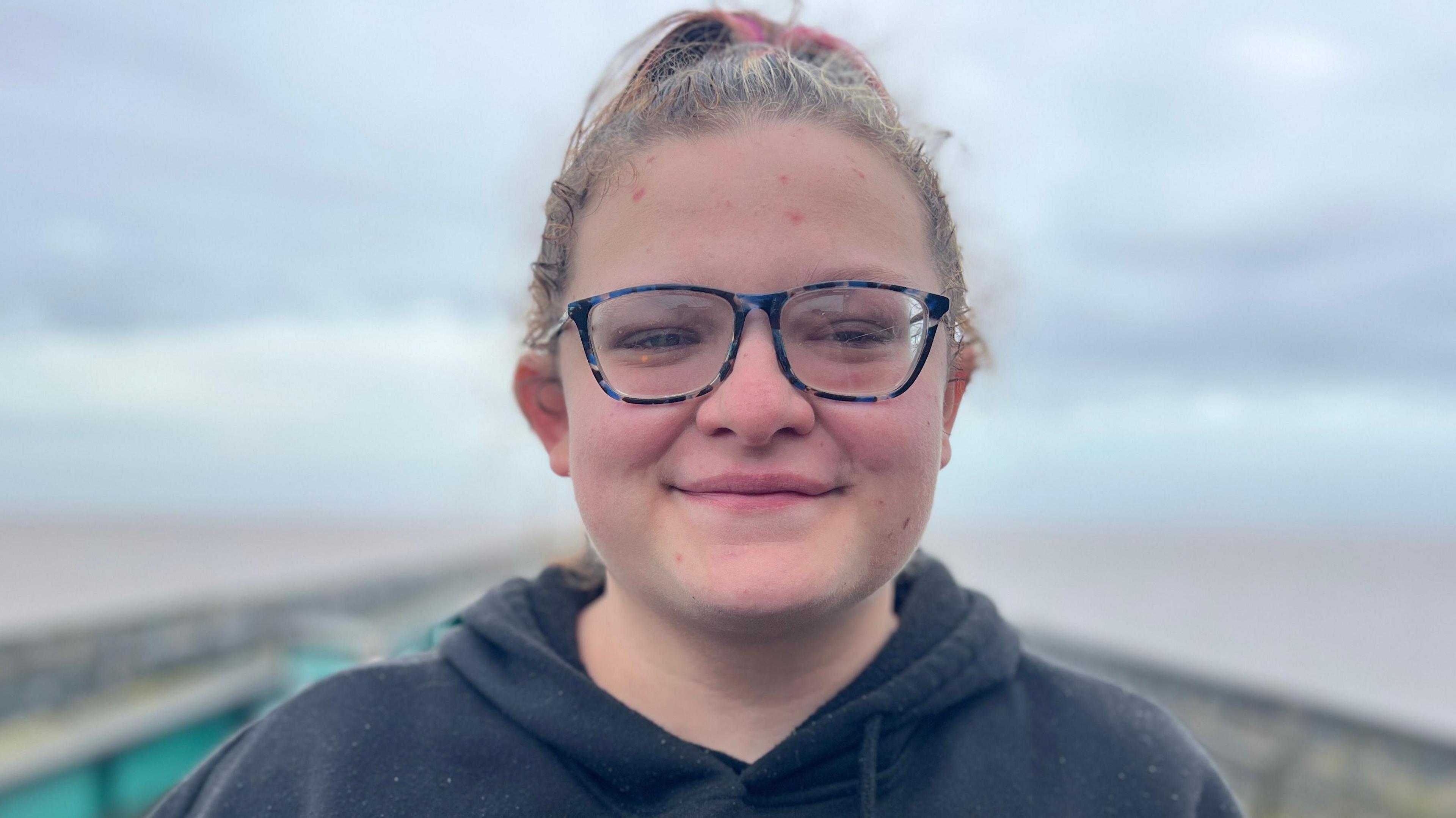 Image of Tasha Stone standing outside on Clevedon Pier. She is wearing glasses and a black hoodie. Her hair is light and tied back. She is looking directly at the camera and smiling. 