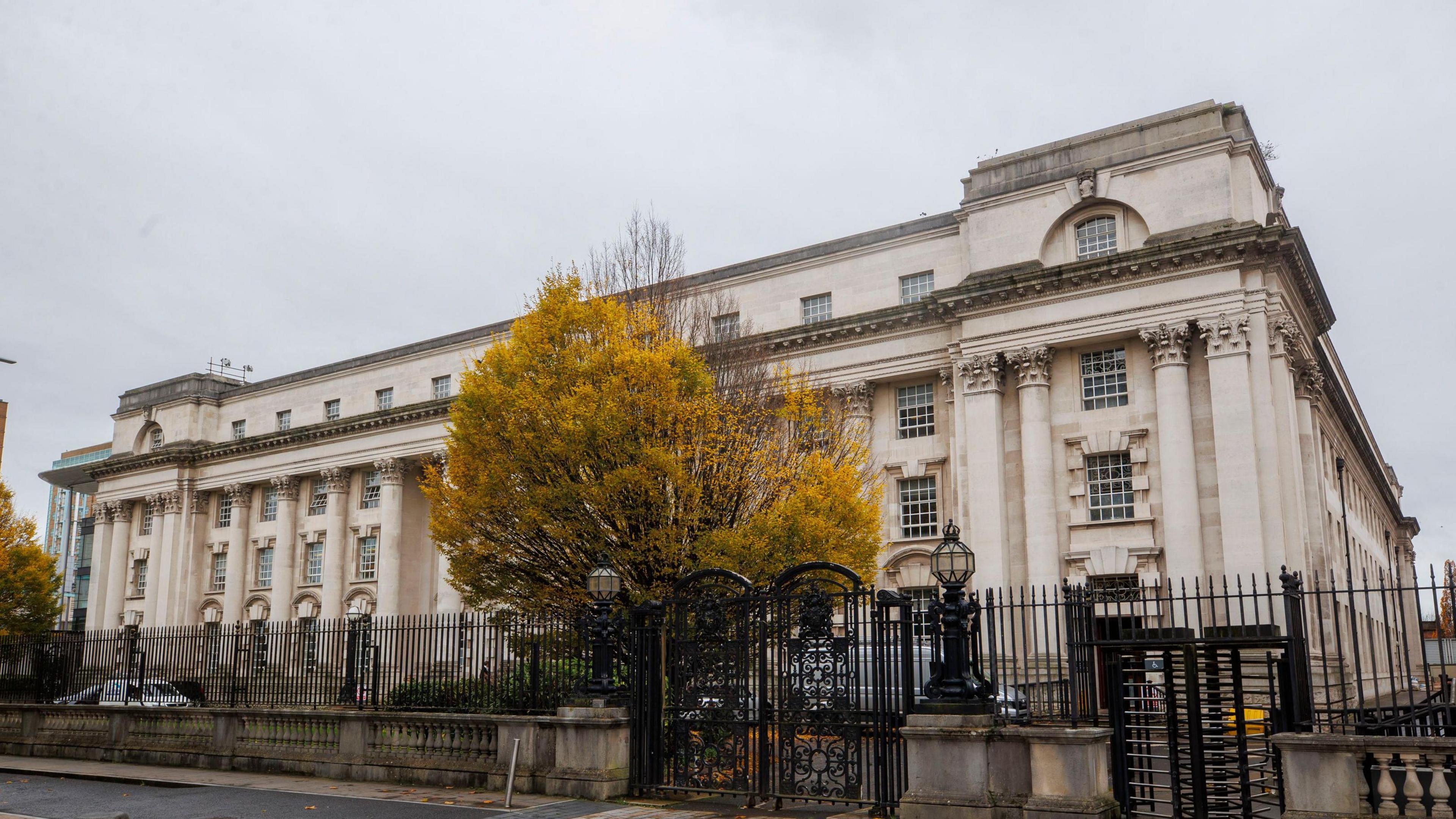 The Belfast High Court is a stone coloured building in Belfast with black metal fences and gates around it. There is a green tree in front of the building.