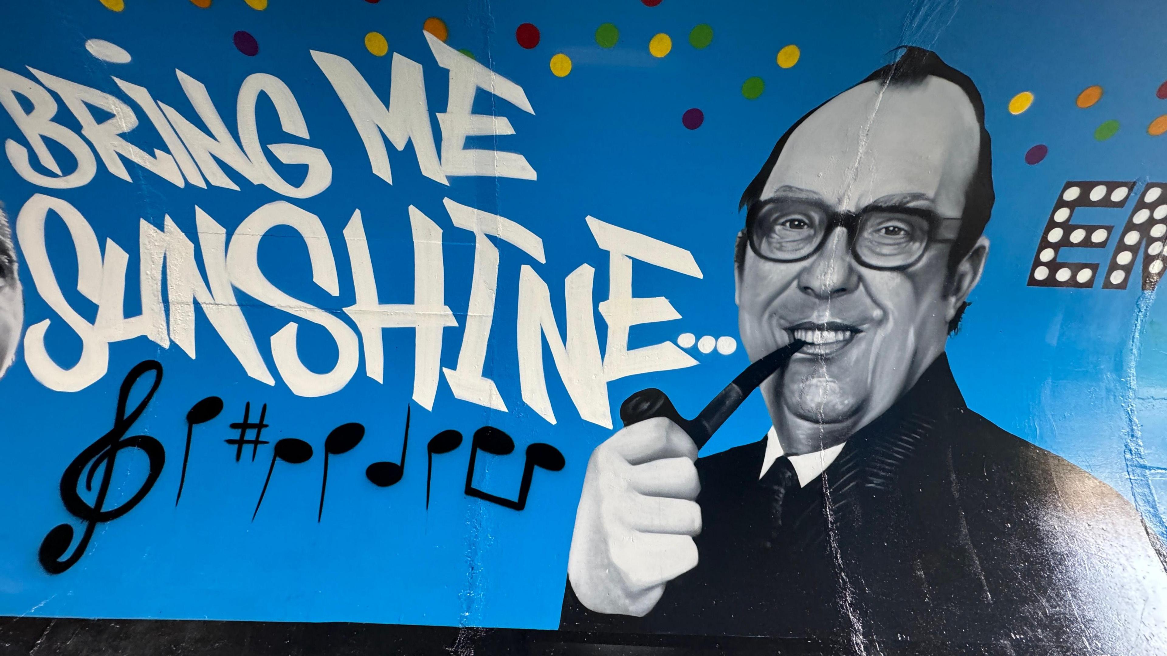 A painting of Eric Morecambe on a wall with lyrics to Bring Me Sunshine and musical notes written next to him