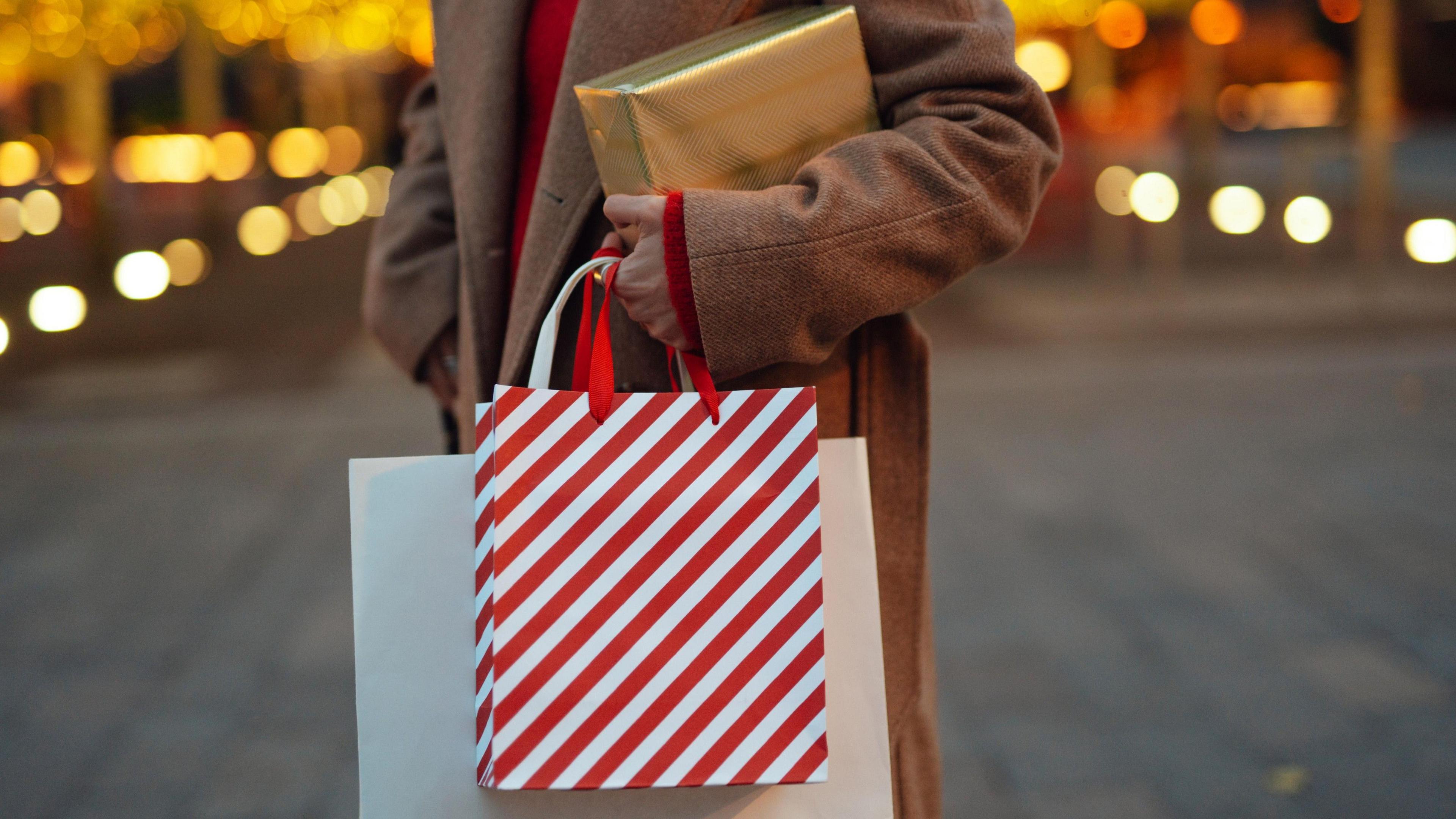 A woman wearing a brown overcoat and red sweater stands in a street decorated for Christmas, holding a gold-wrapped parcel and two shopping bags.