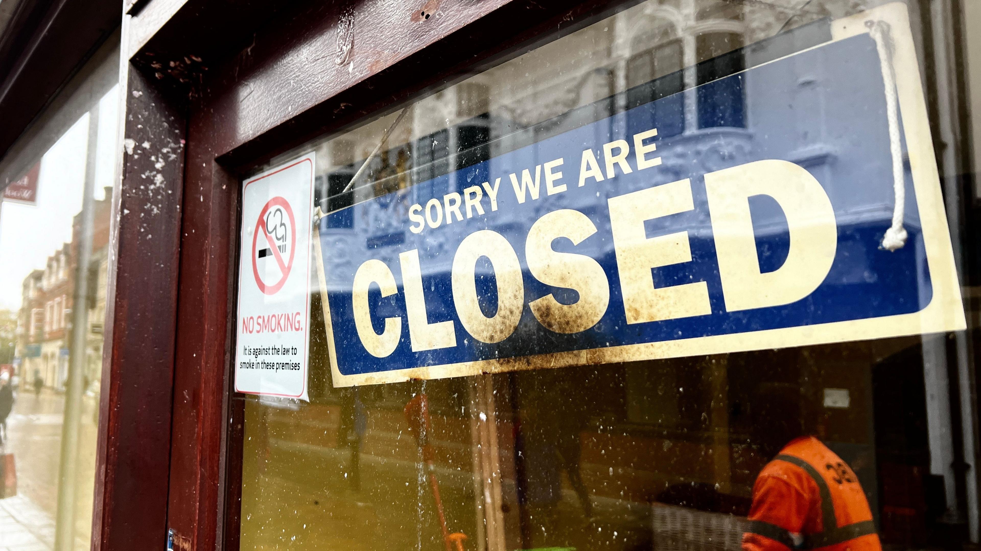 A sign on a closed shop front reads, "Sorry we are closed". The front's glass is tinged from dirt and shoppers can be seen in its reflection walking past.