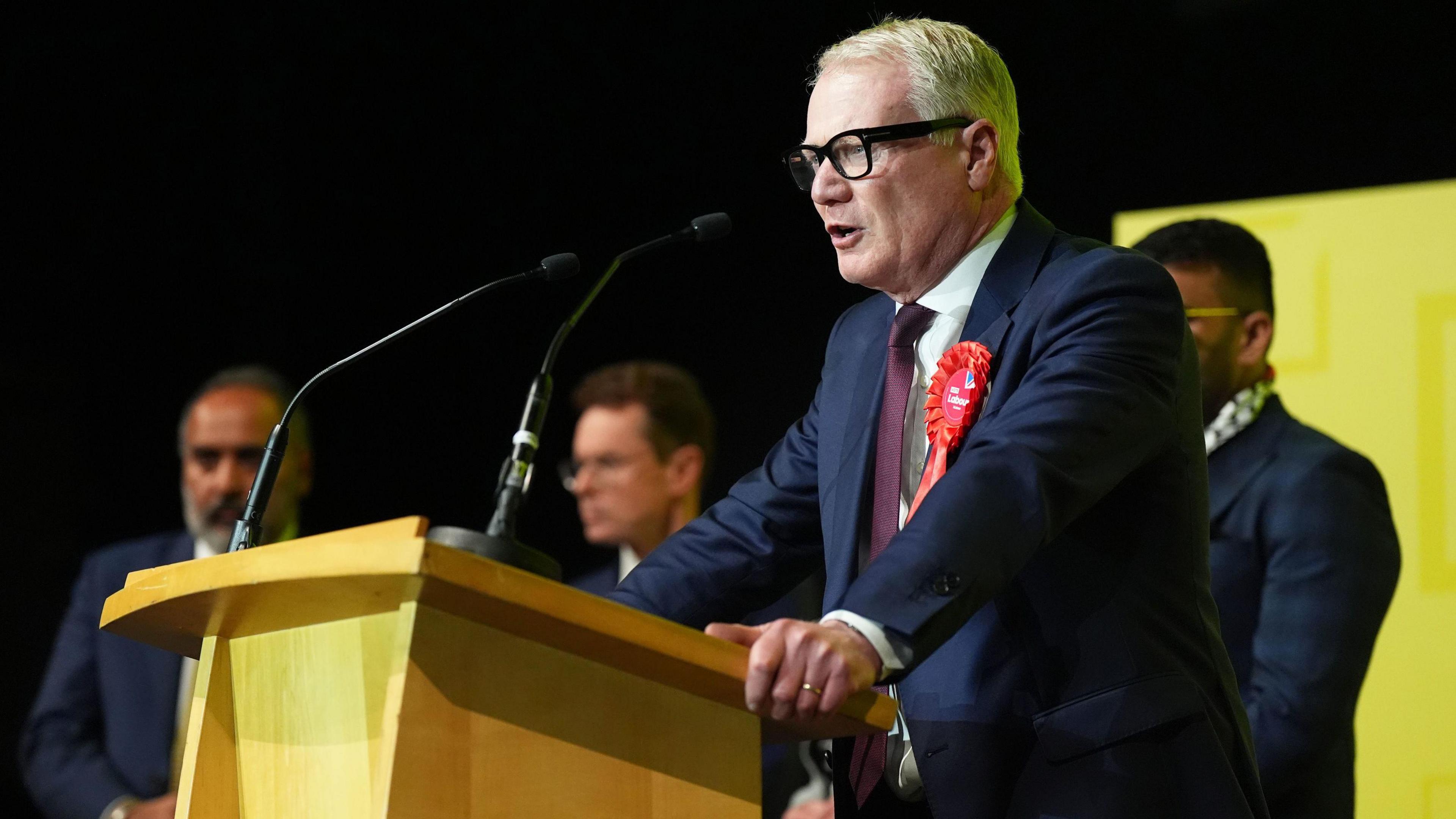 Richard Parker - a man with short white hair, glasses and wearing a suit with a red rosette - stands at a podium speaking into a microphone as three men stand behind him. 