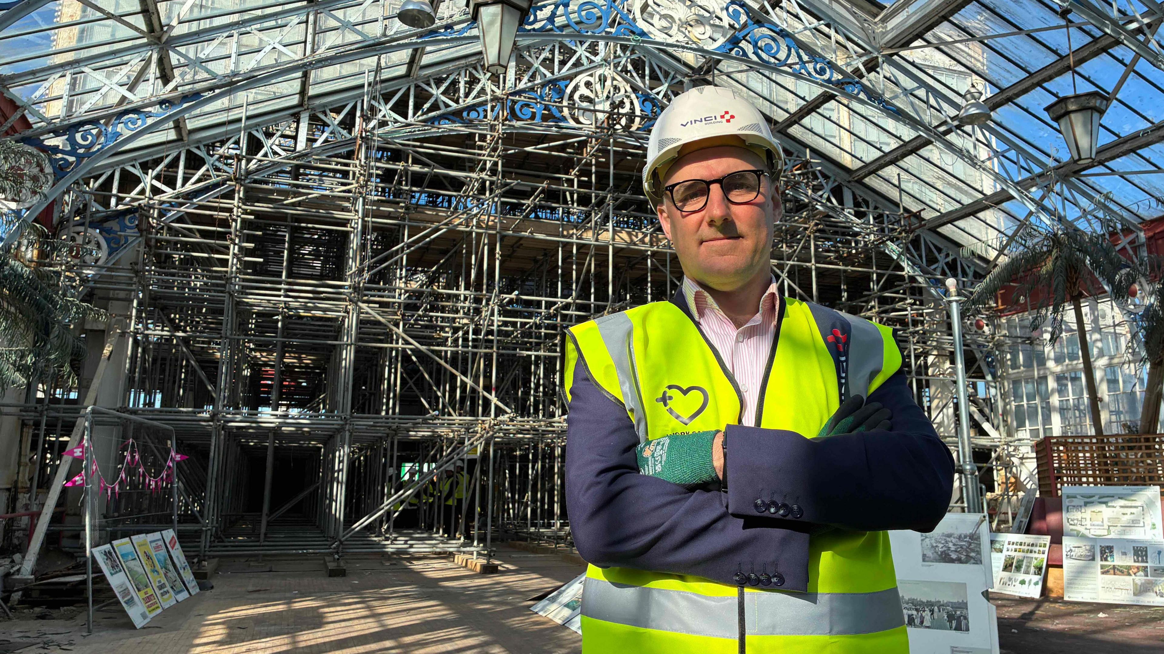 Paul Tumelty standing in a blue suit and pink shirt with his arms crossed. wearing a high vis standing in the Winter Gardens behind him all the scaffolding on show