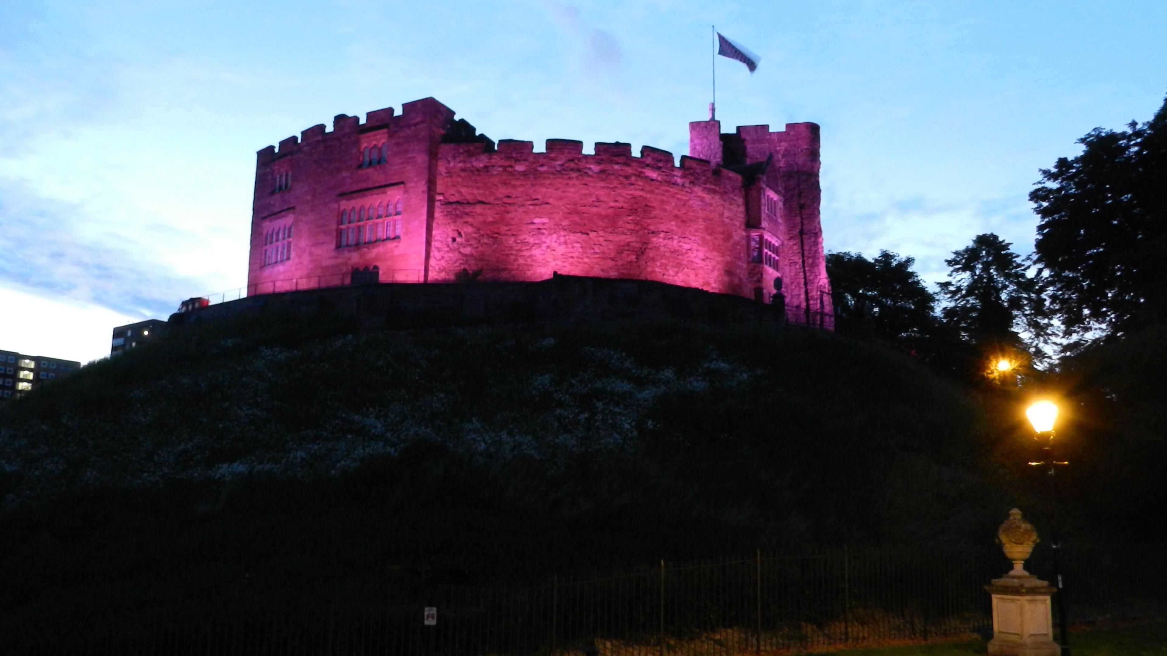 Tamworth Castle lit up in purple at twilight, with a flag flying. There are lamps lit in the street below.