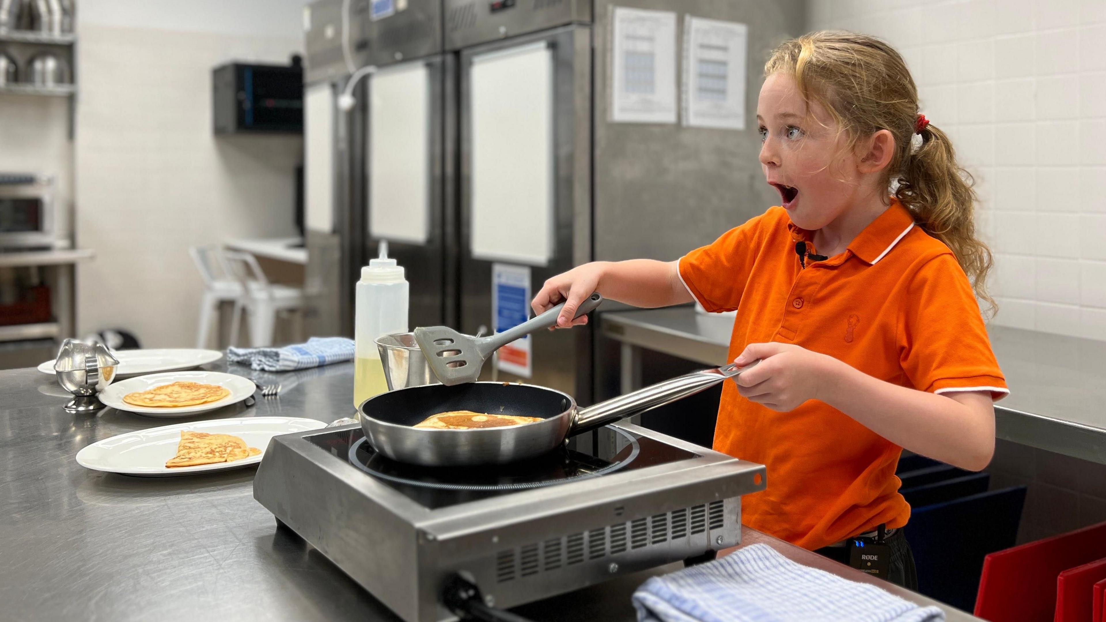 Ophelia is in an orange shirt cooking pancakes with a spatula in a commercial kitchen, surrounded by stainless steel appliances, metal bowls, and plates of pancakes on the counter