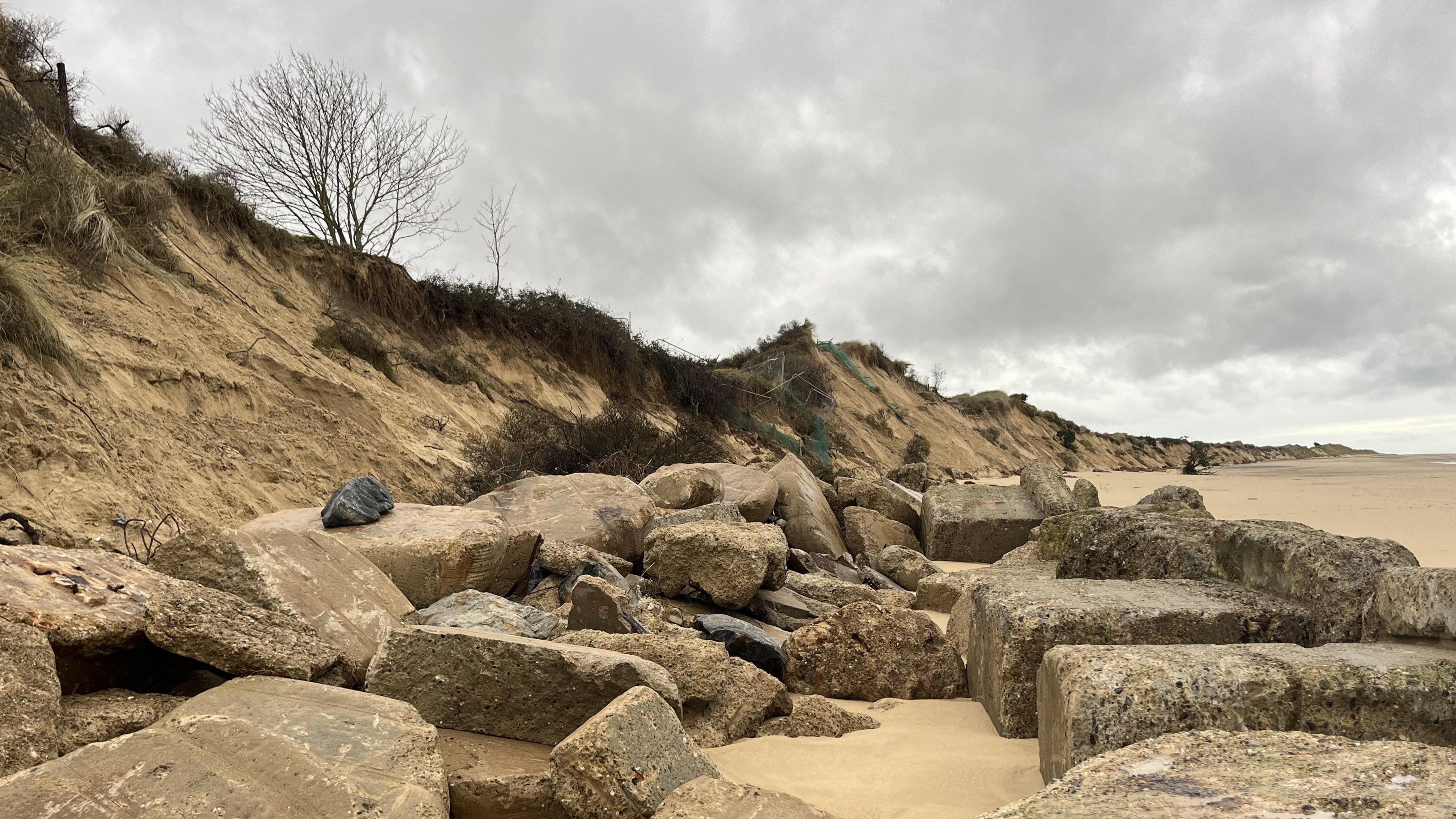 Large sea rocks line a sandbank on a beach. Greenery and shrubs line the bank while a tree can be seen further up. It is a grey cloudy day.