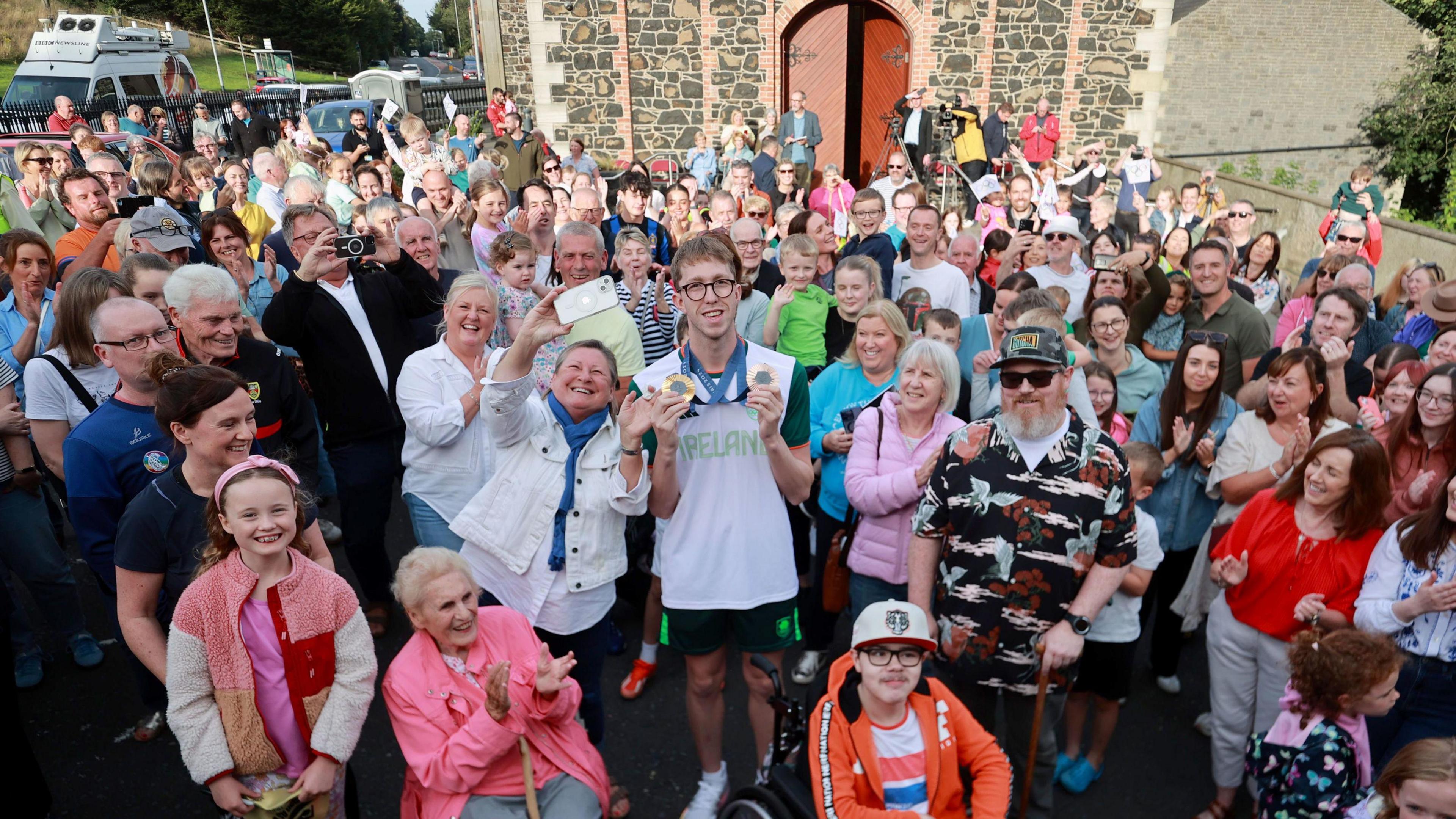 Daniel Wiffen is standing in the middle of a large crowd of people in Magheralin. He's wearing his Team Ireland top and shorts and holding up his gold and bronze medals.