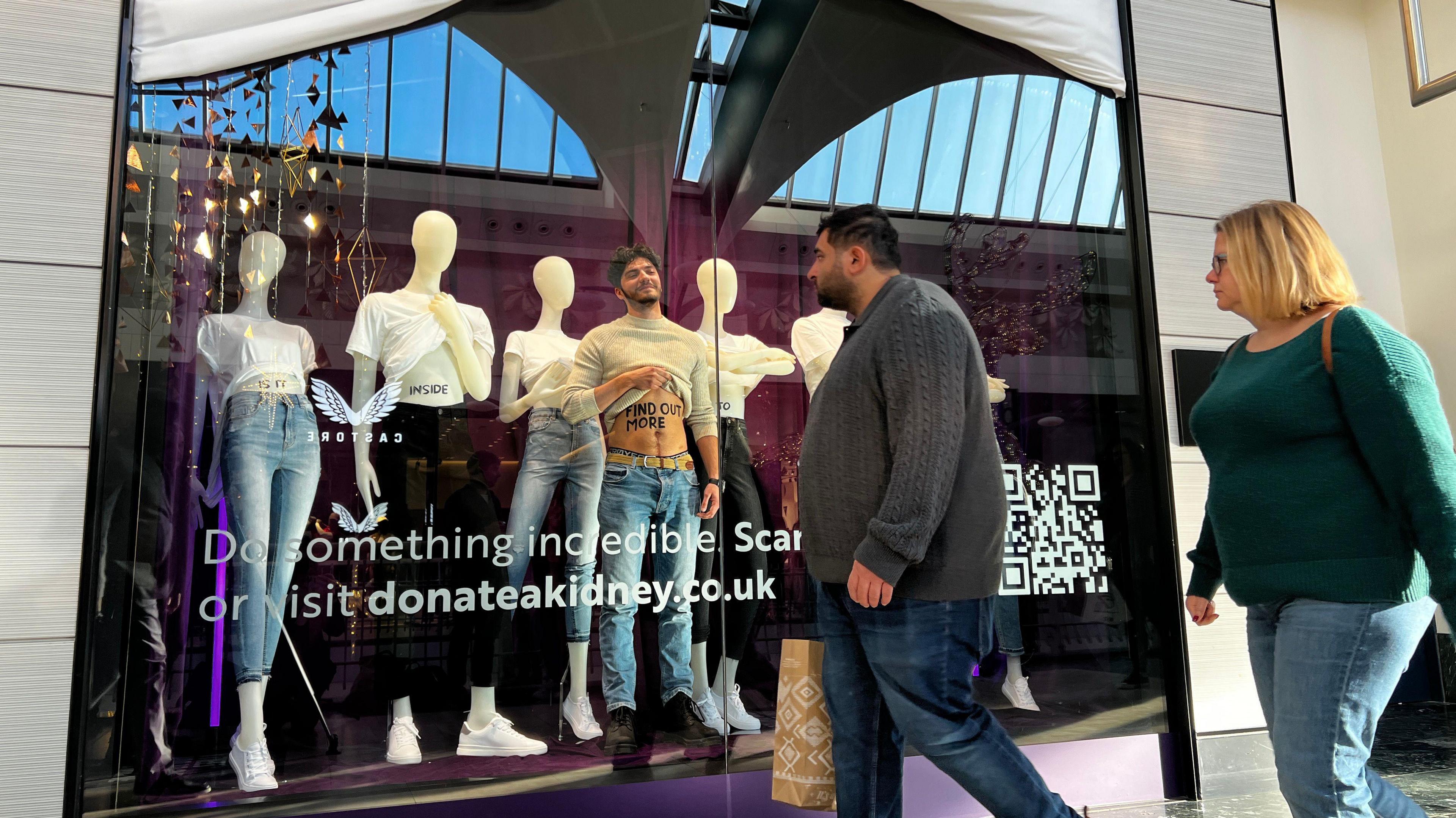 George Cannon stands in a window in Bluewater Shopping Centre, raising his shirt as part of a campaign to encourage more organ donation. He stands in front of six mannequins, as a male and female shopper look on as they walk past.