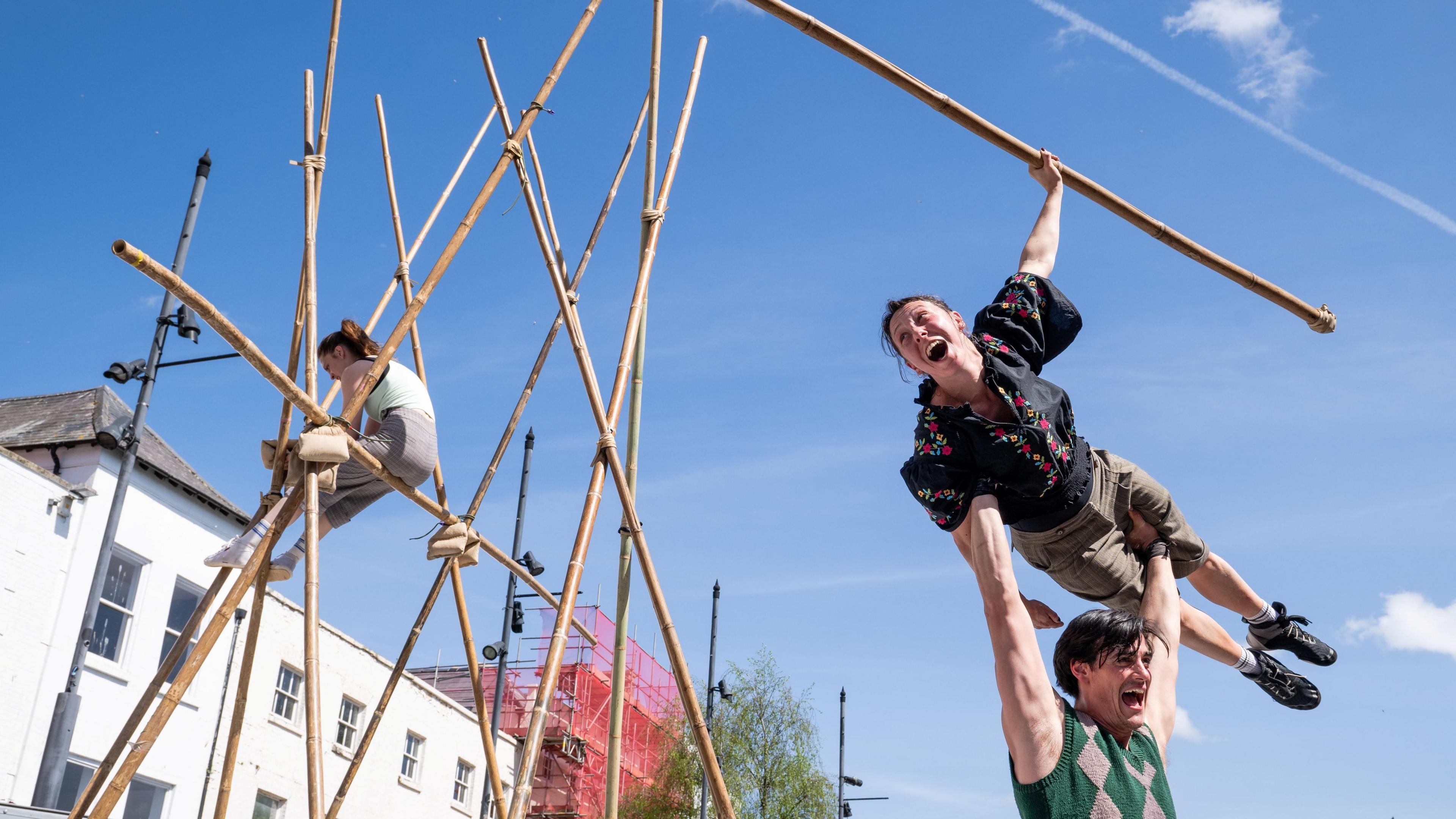 Three performers are posing amid a series of bamboo poles. One of the performers is holding up another who is horizontal and holding a pole aloft. A third is sat, perched on poles nearby.