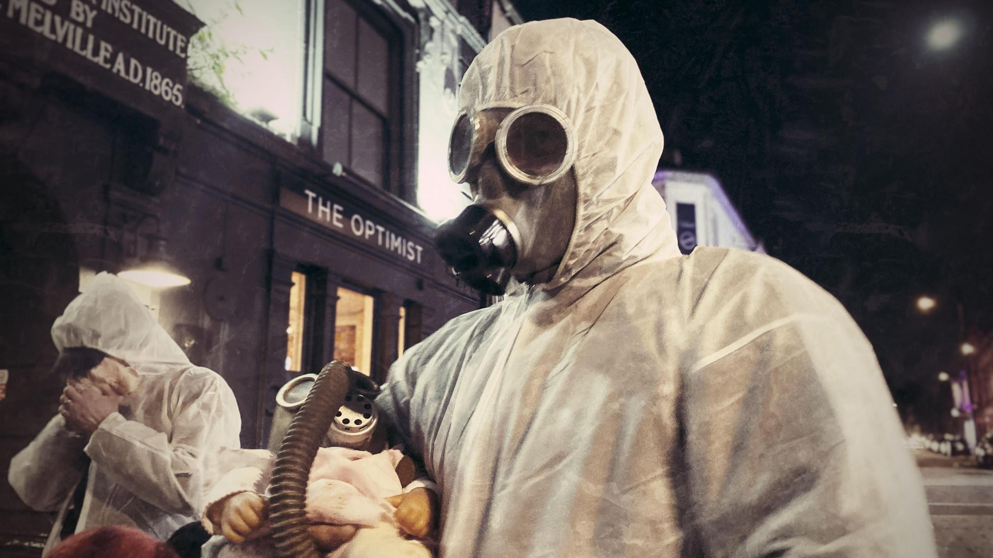 Two people dressed in white personal protection equipment that covers their bodies and wearing gas masks. It is night, and they are passing a pub called "The Optimist" in a street in Northampton.