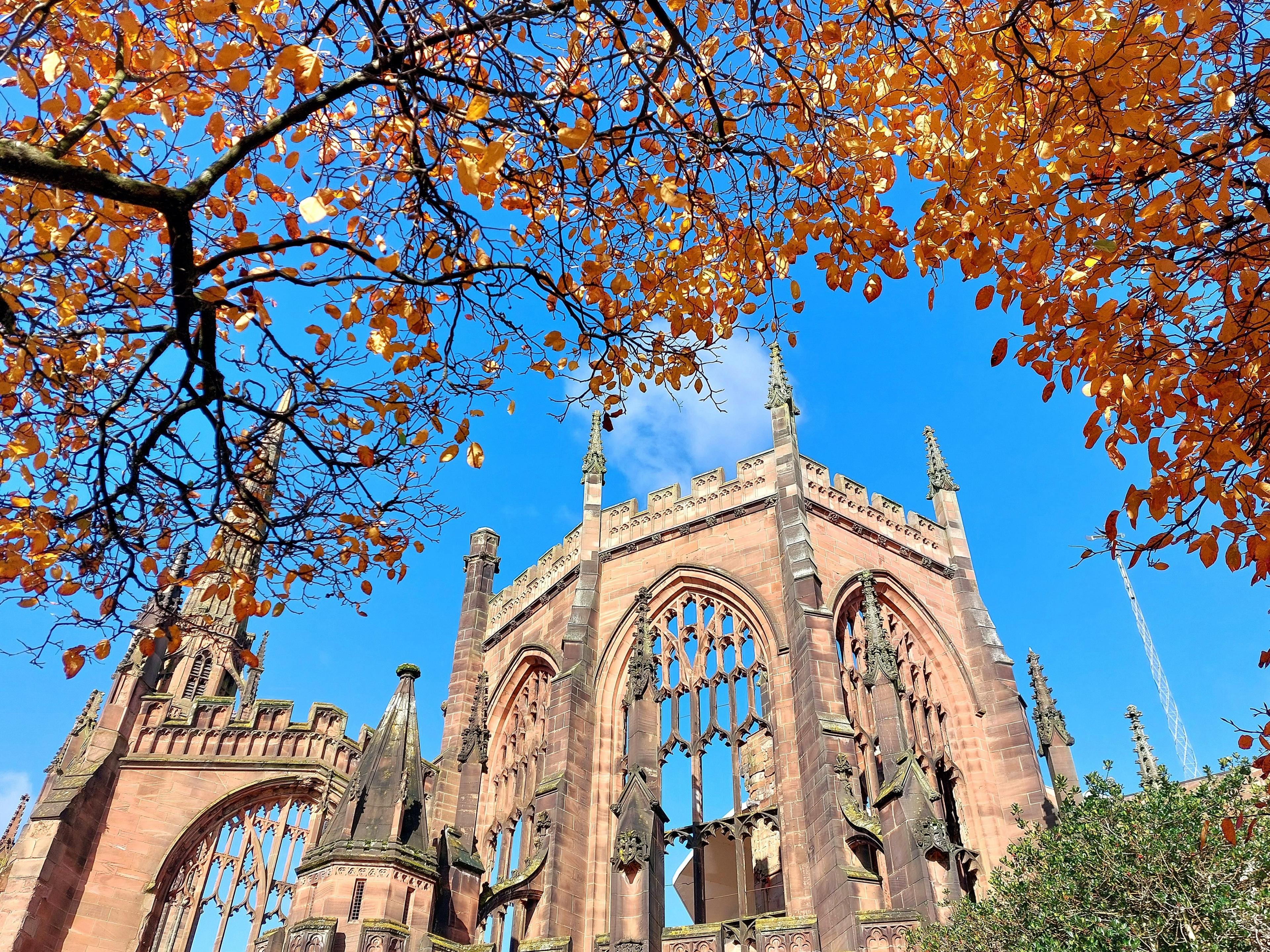 Roofless ruins of the cathedral with blue sky showing through the windows and above. Yellowing autumn leaves hang from a tree in the foreground.