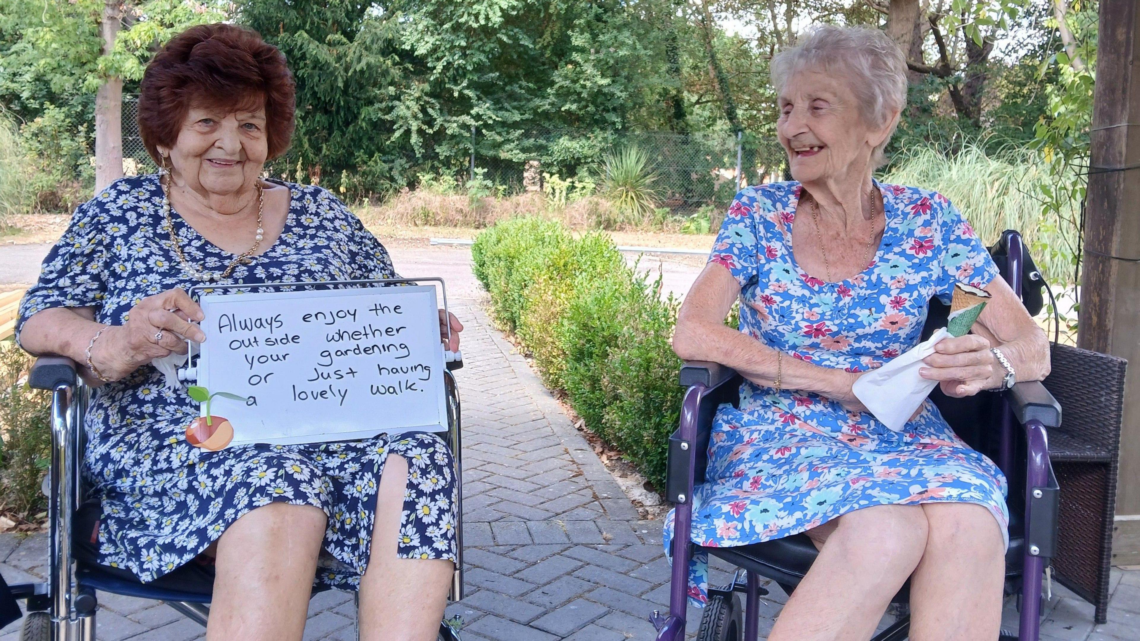 Two women in flowery dresses sitting in wheelchairs. The one on the left has short brown hair and is holding up a whiteboard saying "always enjoy the outside whether you're gardening or just having a lovely walk", and smiling at the camera. The woman on the right is looking at the woman on the left and smiling.