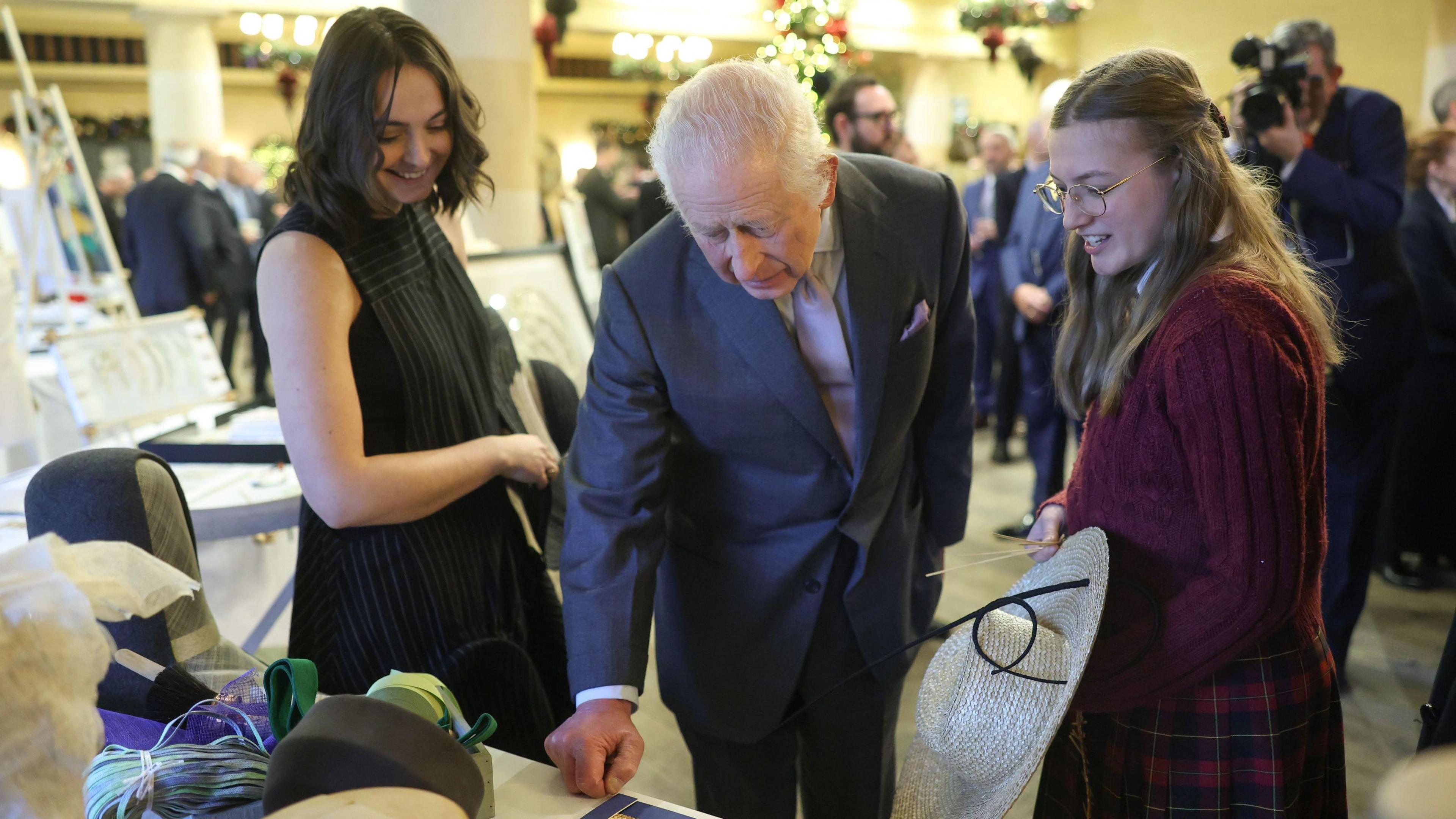 King Charles III pictured next to two young women. The one to the left of the frame has short dark hair and is wearing black. The woman on the right has long blonde hair, glasses, and is wearing a red dress. King Charles is wearing a dark grey suit jacket and trousers with a white collar shirt and pink tie. All three of them are standing at a table covered with crafts and hats. None of them are looking at the camera - they are looking down at the table.