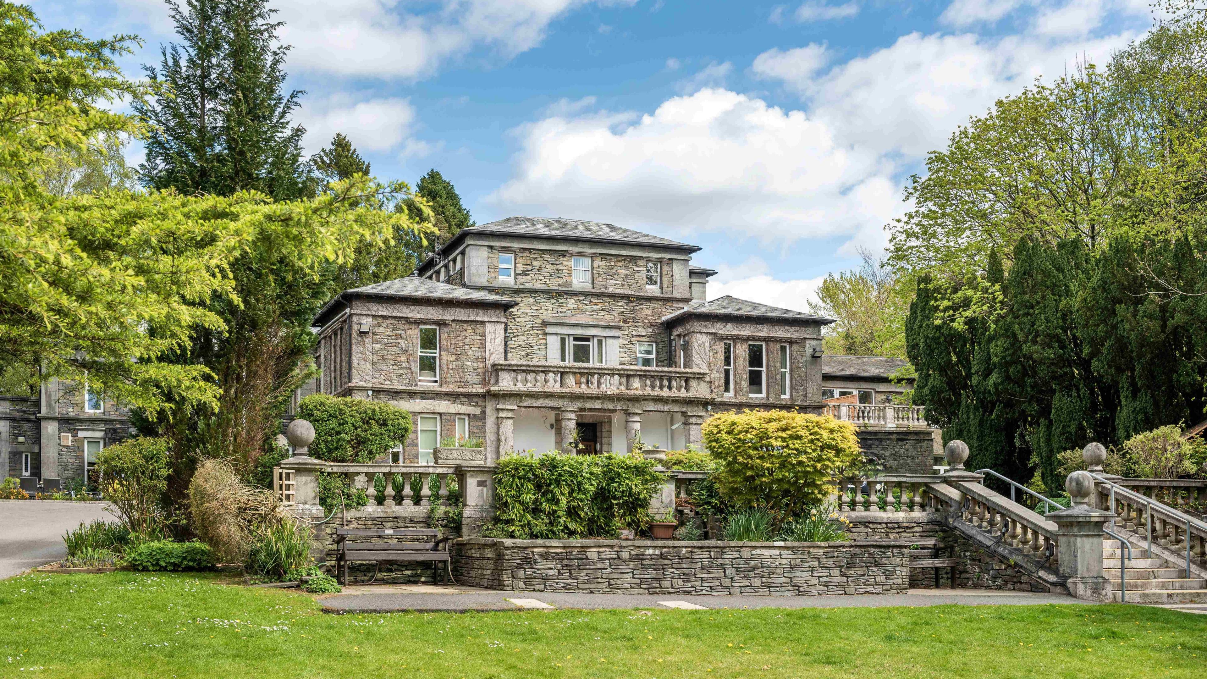 The Windermere Manor Hotel. It is an 1850s Victorian residence with a balcony and terrace at the front and a manicured lawn.