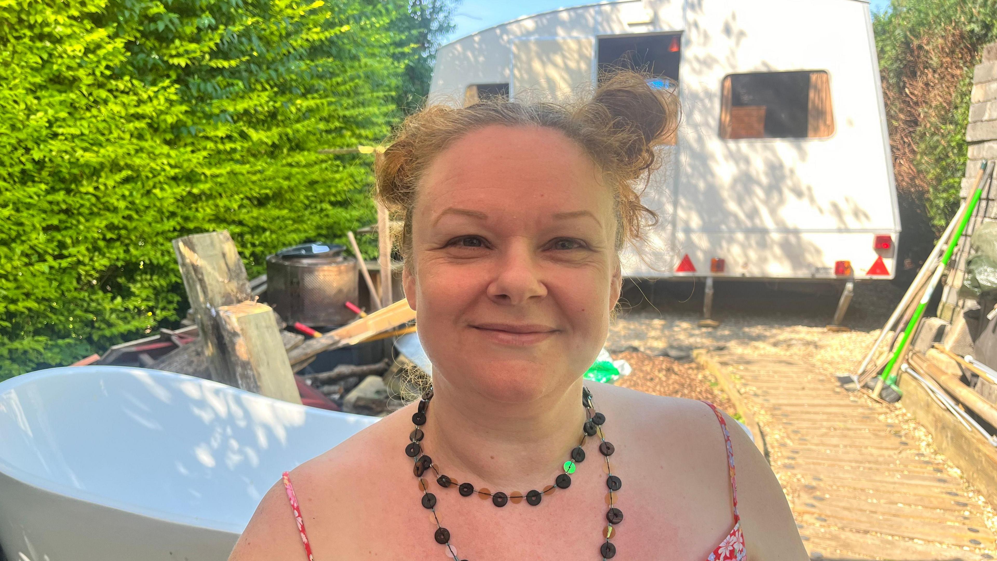 Adele is smiling and standing in front of the pop-up caravan in her garden. There is a privet hedge on the left and a bathtub.