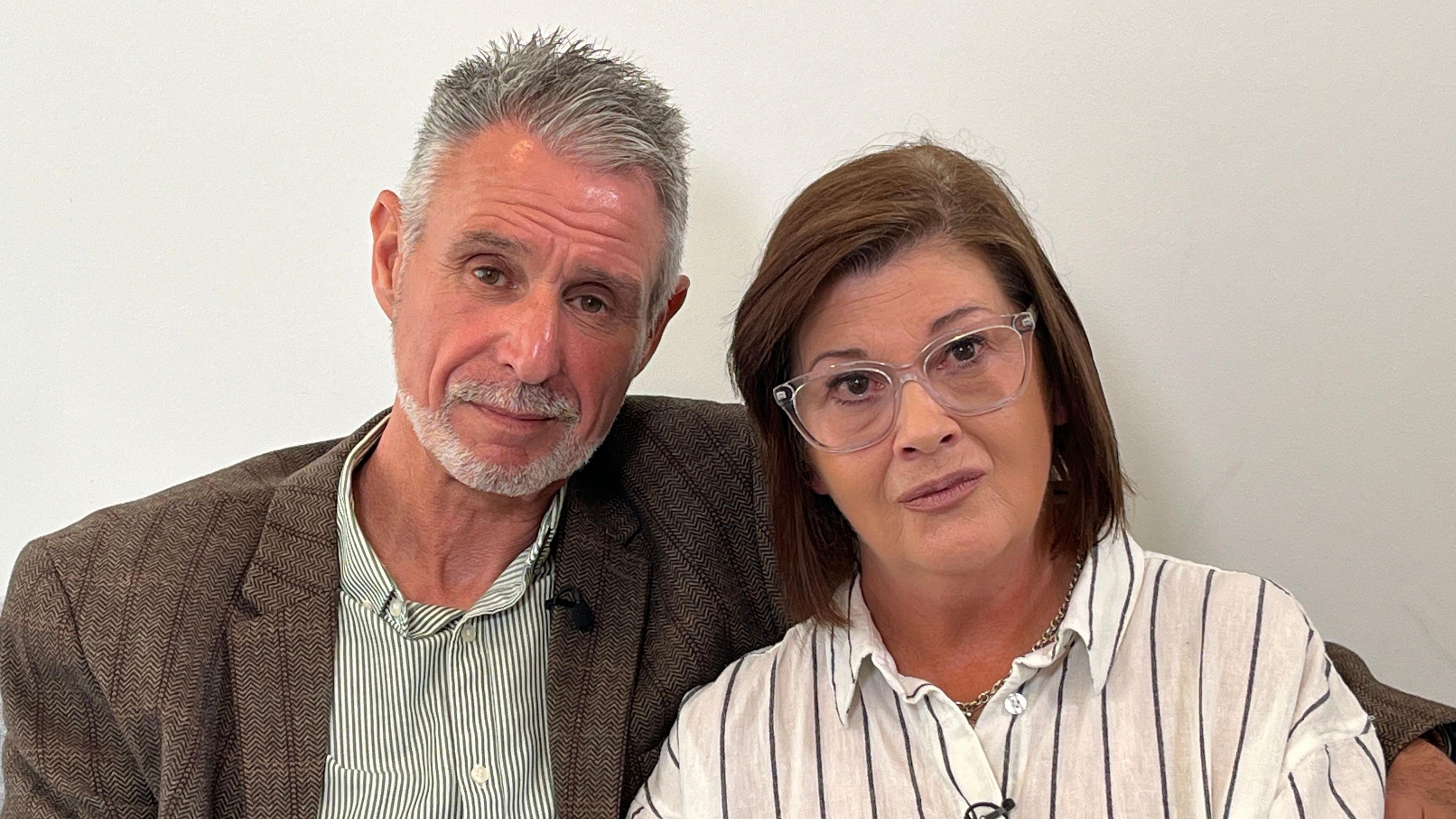On the left Andrew Bashford has short grey hair with a grey goatee beard. He wears a brown checked jacket over a green striped shirt. On the right Sharon Bashford has shoulder-length dark brown hair. She wears glasses and a white shirt with black stripes.