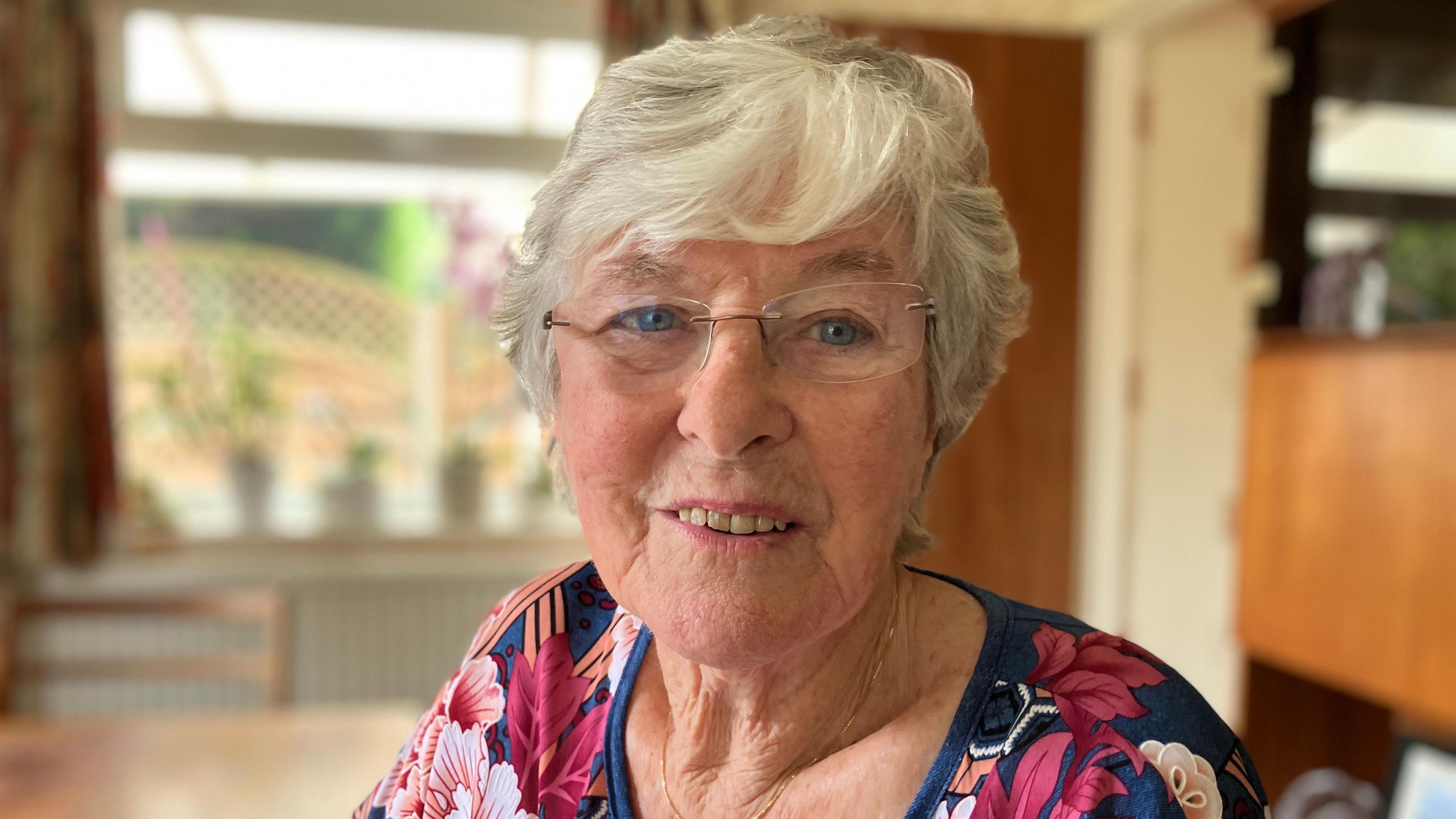 A smiling Maureen Bedford looks directly at the camera as she is photographed in a dining room. She is wearing a blue top with a flowery print, glasses and has short grey hair. 