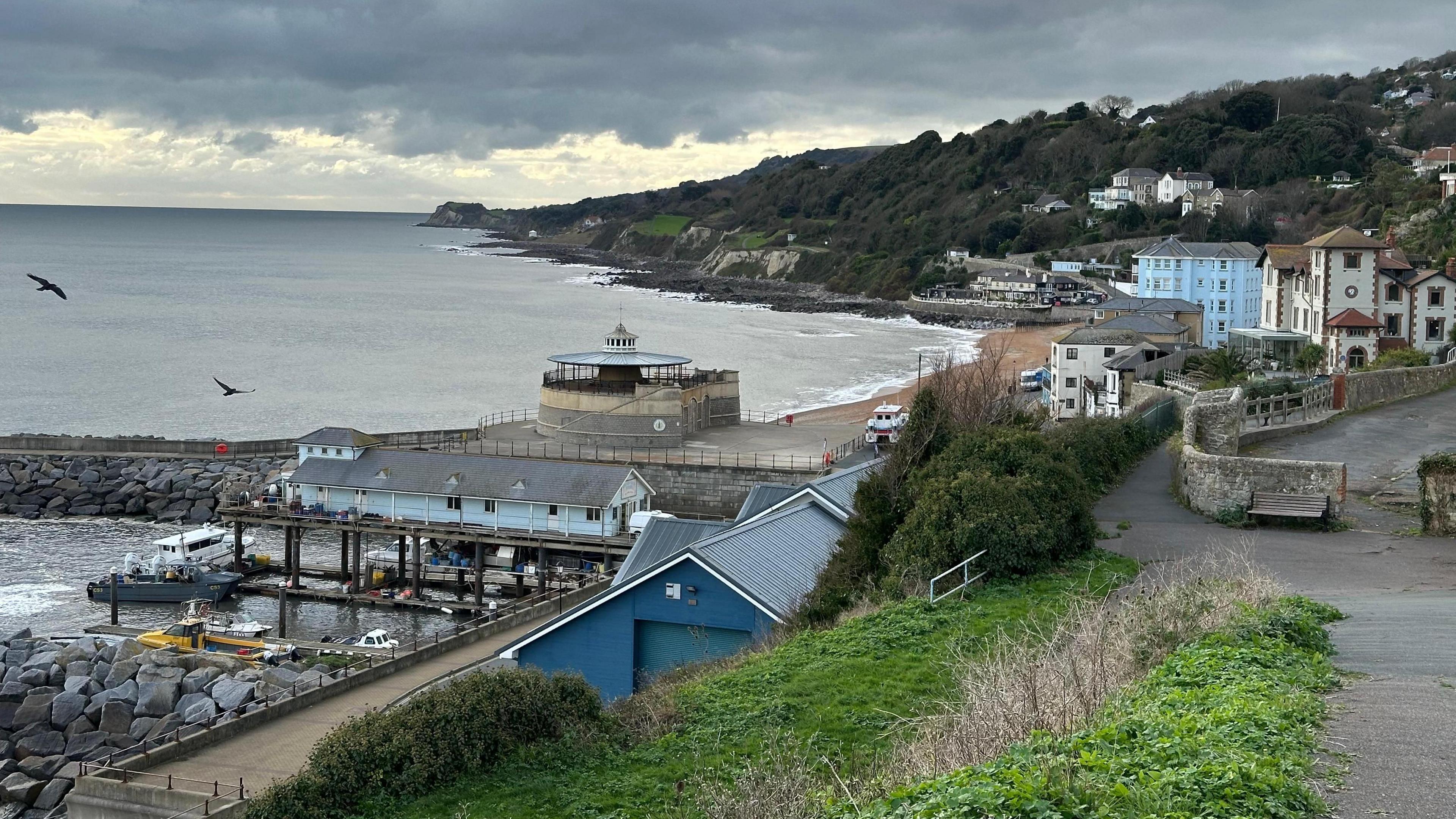 An aerial view of the beach at Ventnor with several buildings in the centre of the picture running alongside the sand. In the distance the coastline turns more rugged with trees and cliffs visible. In the foreground, there are sea defences made up of large boulders.