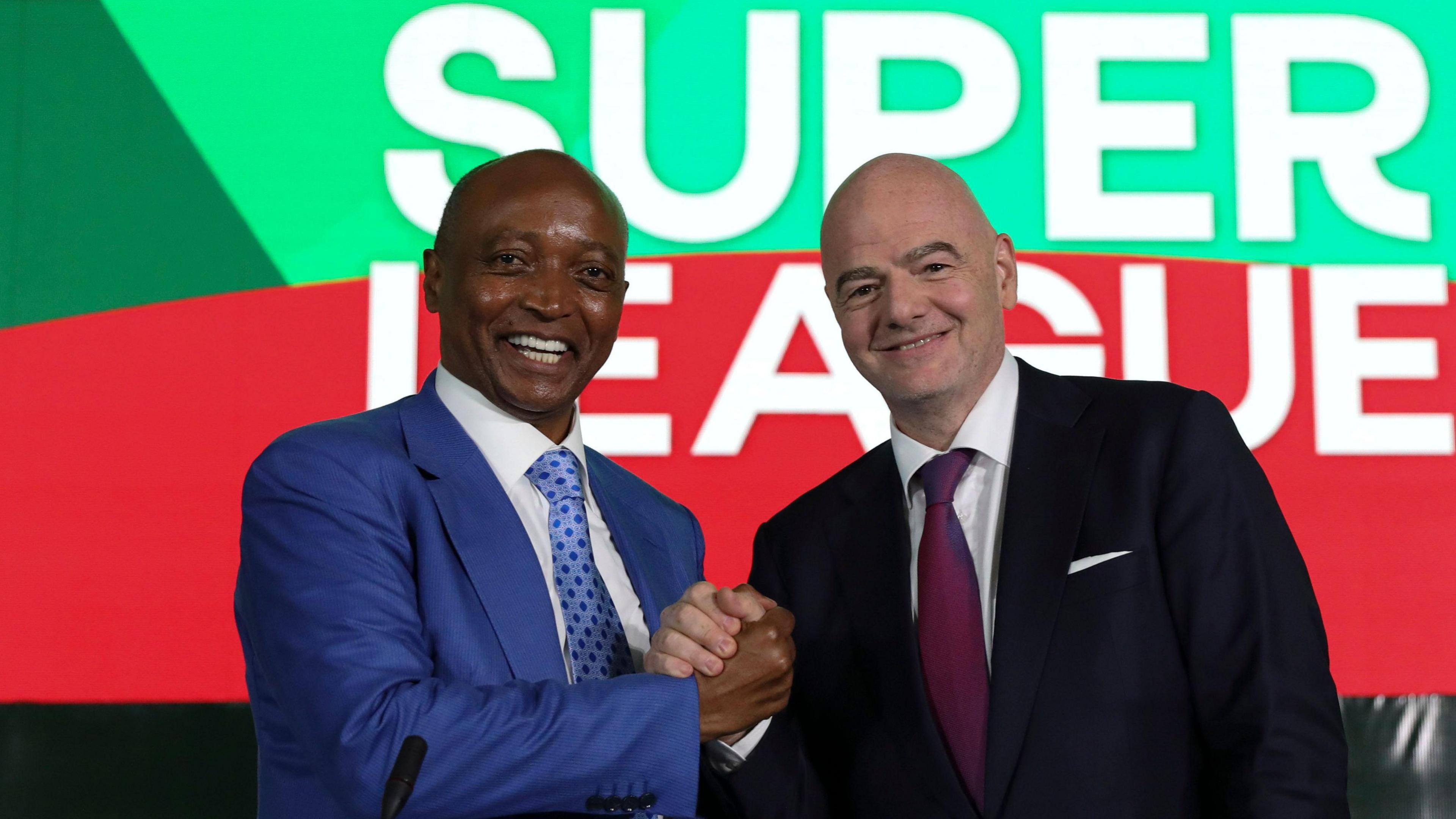 Patrice Motsepe, wearing a blue suit, white shirt and blue patterned tie, clasps hands with Gianni Infantino, who is wearing a black suit jacket, white shirt and purple tie. The two men, visible from stomach up, smile as they look directly into the camera, with the wording 'Super League' visible on a wall behind them