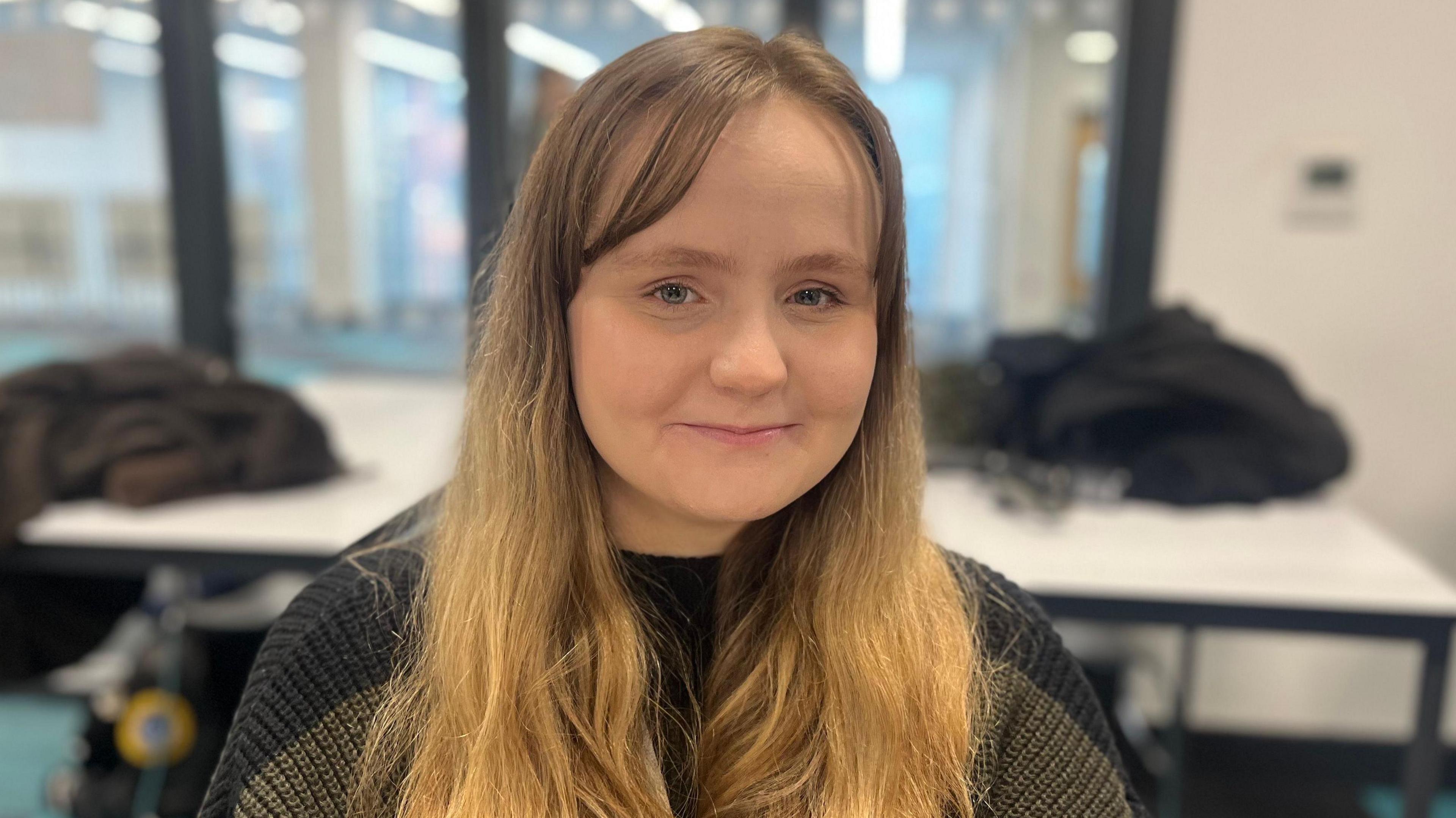 Head and shoulders of Evie Whitehead who is wearing a black jumper. A table with coats and bags on can be seen behind her.