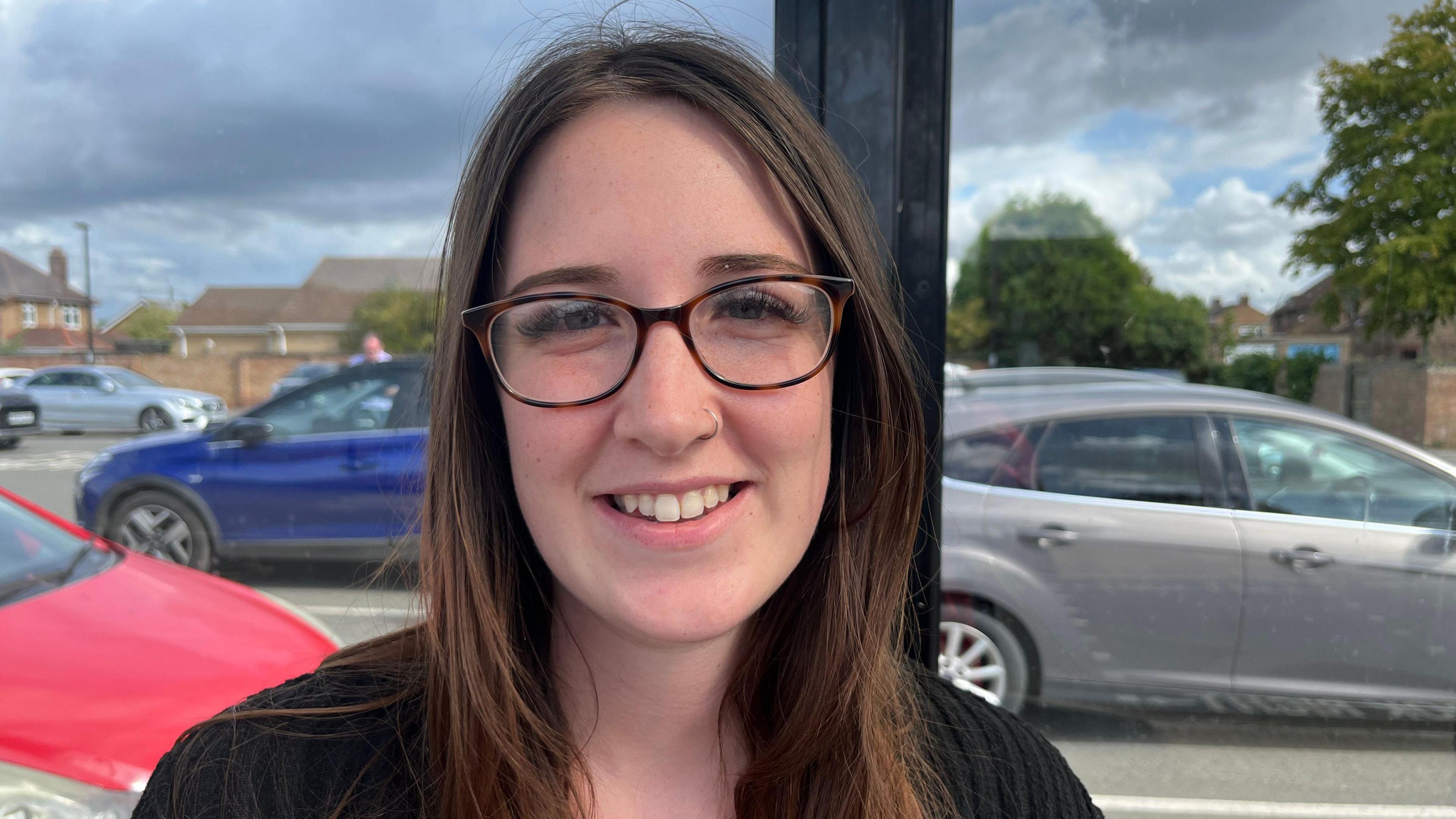 Ms Merry is smiling at the camera while standing outside at a bus stop that backs onto a car park. She has long brown hair, circle framed dark glasses and is wearing a black top.