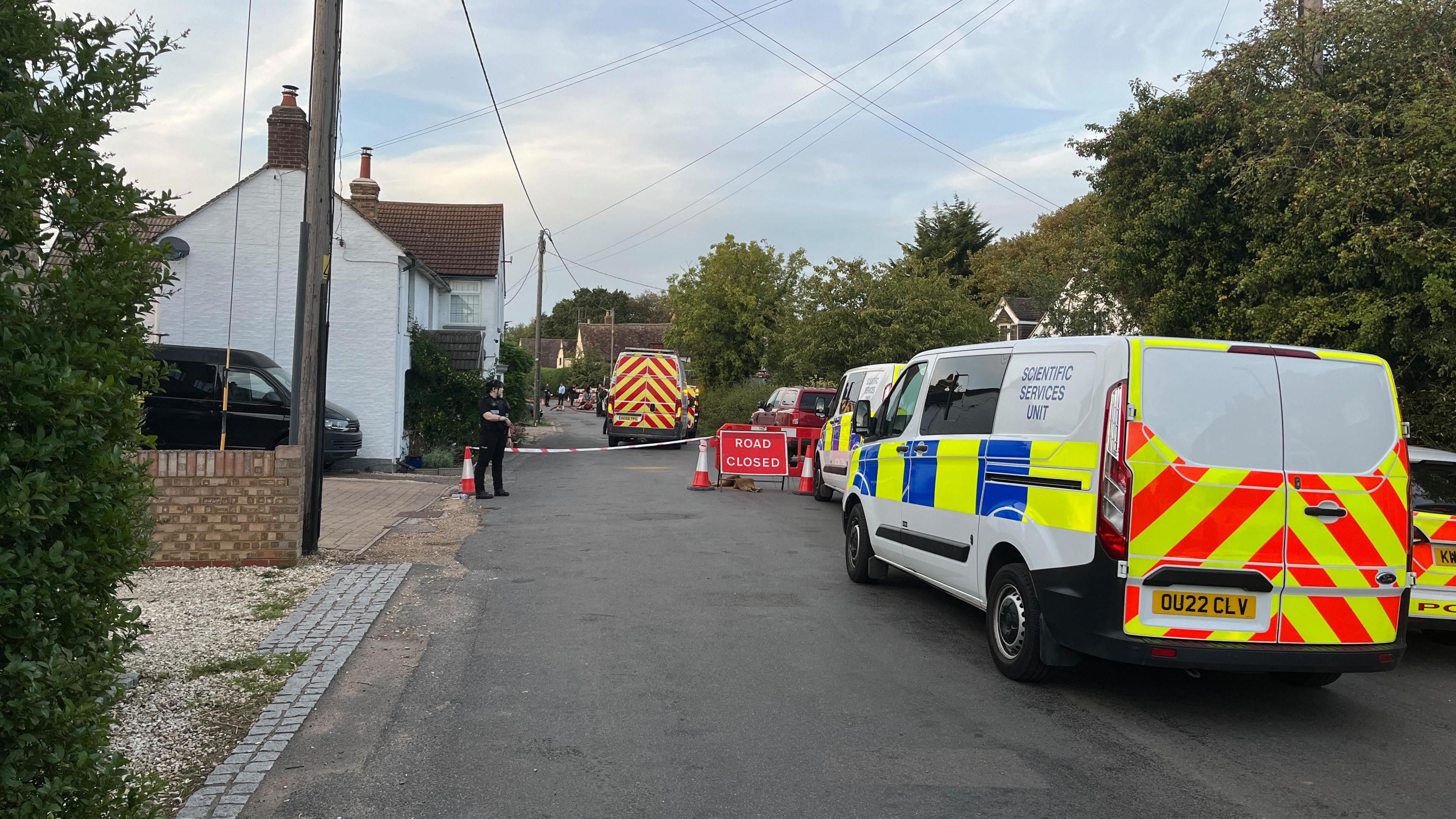 Police scientific vans on a road which has a "road closed" sign on it. There is a female police officer standing by a cordon. There are houses on either side of the road.