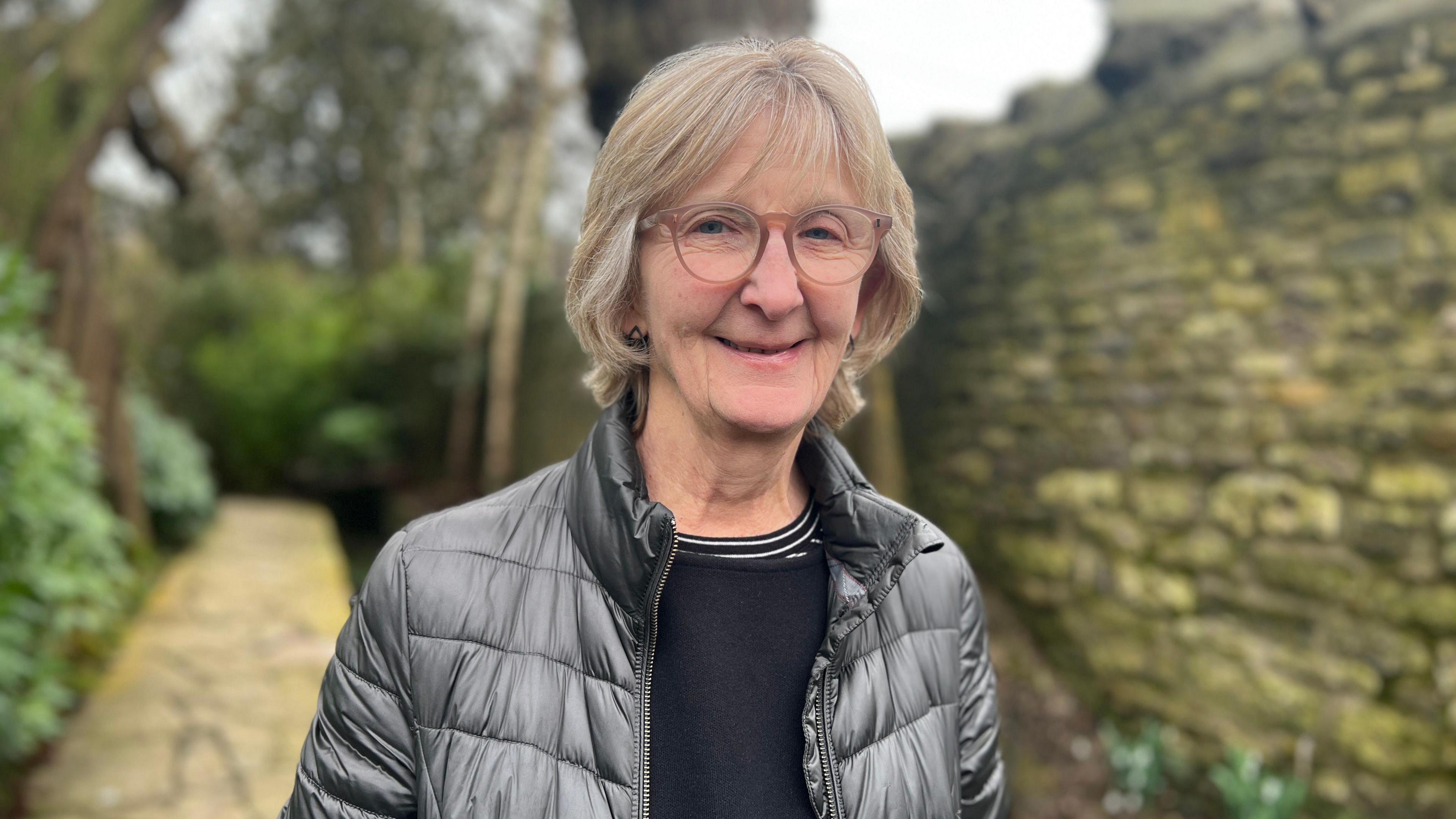 Amanda Hirst is standing in front of a garden path and a stone wall. She is wearing a grey jacket and is smiling at the camera.