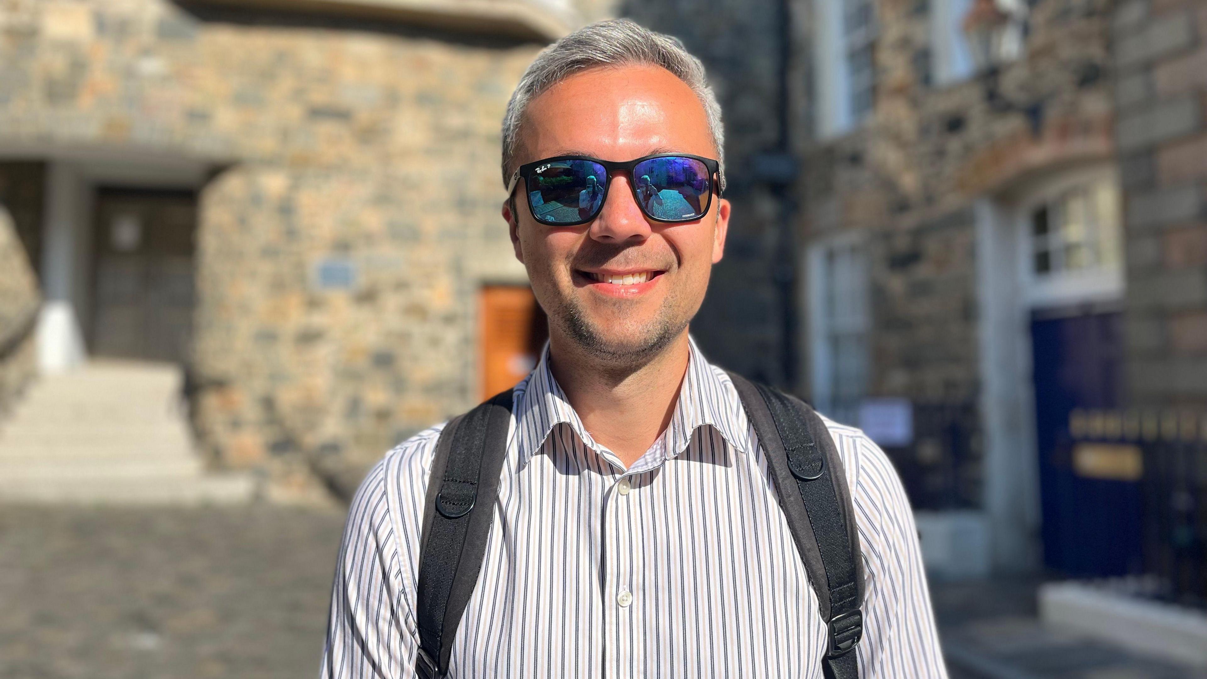 A man with light grey hair smiling at the camera. He is wearing sunglasses and has a pale pink shirt with blue pinstripes. You can see the straps of a grey rucksack over his solders.