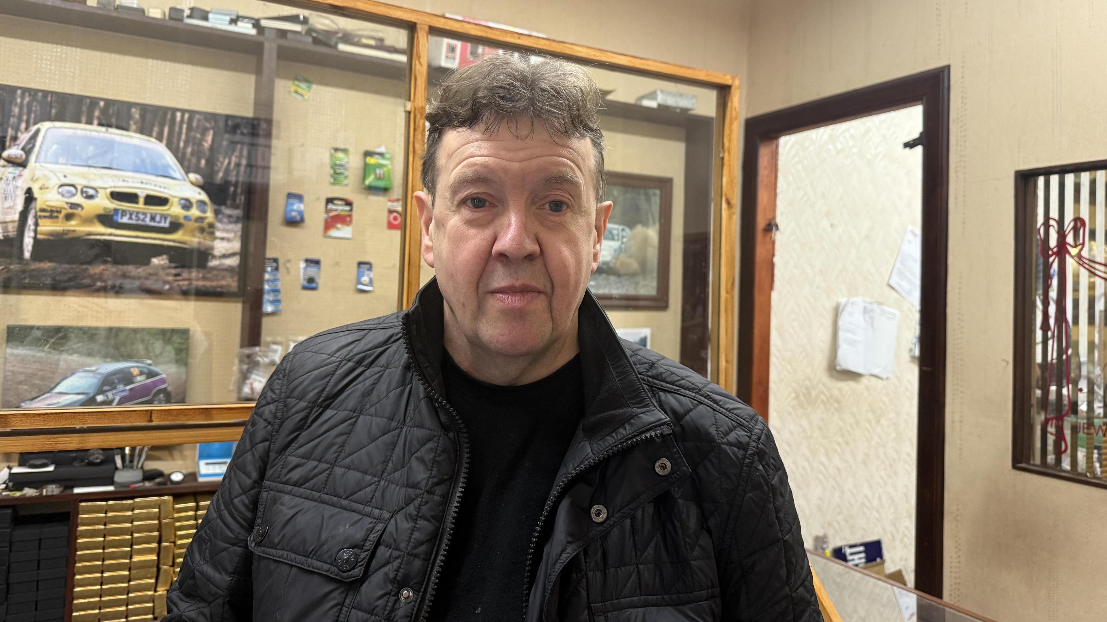 A man with a black coat, black t-shirt, and short brown hair stands in a shop. There is a picture of a yellow car on the wall behind him.