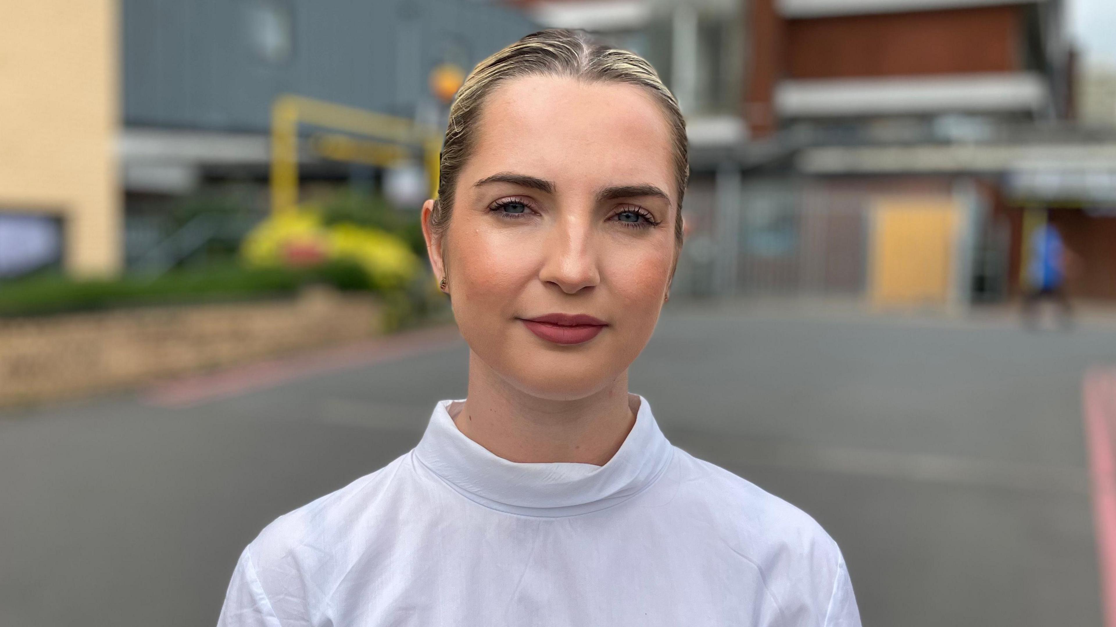 Rosie Wrighting looking at the camera with her blonde hair scraped back in a ponytail. She is wearing a white long sleeved top. 