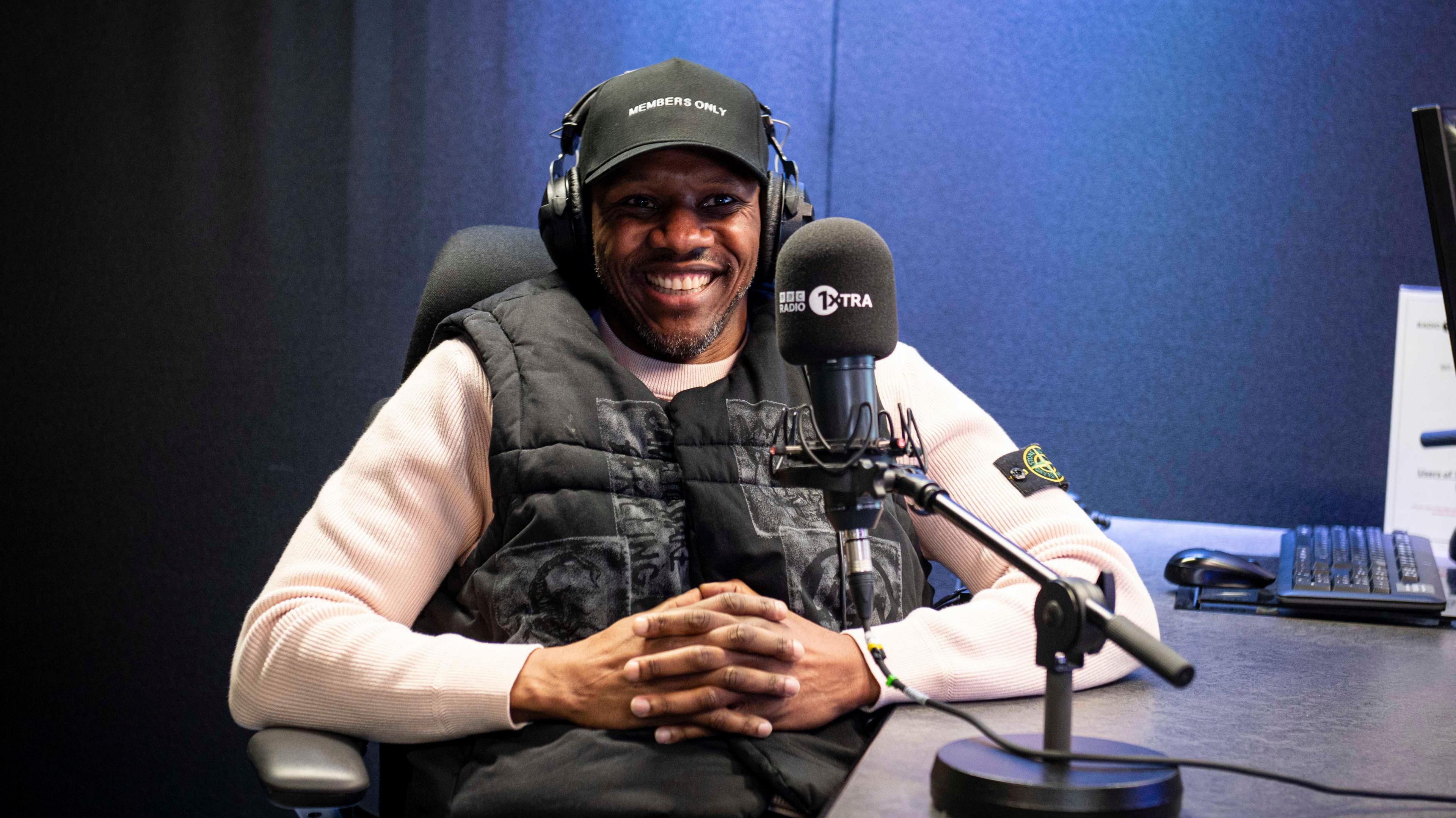 Rapper Giggs is sat smiling in a BBC 1Xtra radio studio.  He is wearing a black cap with a black body warmer and white jumper with a microphone in front of him. 