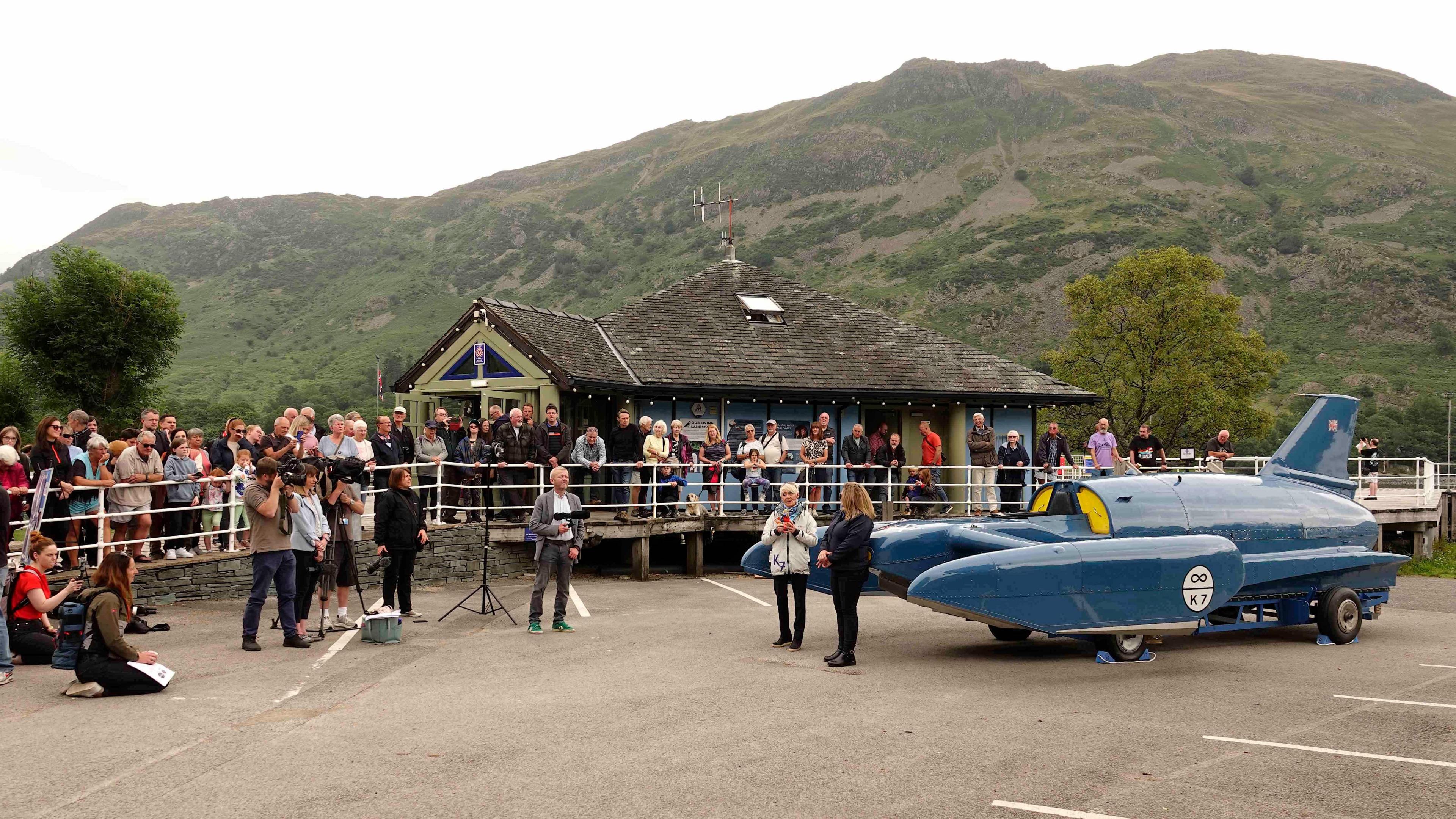 Gina Campbell and Rachel Bell, head of marketing and business development at Ullswater Steamers and Cumbria Tourism’s vice-chair, stand next to Bluebird - a jet-powered hydroplane - in the car park at Glenridding Pier. There are dozens of onlookers and fells can be seen towering above in the background.