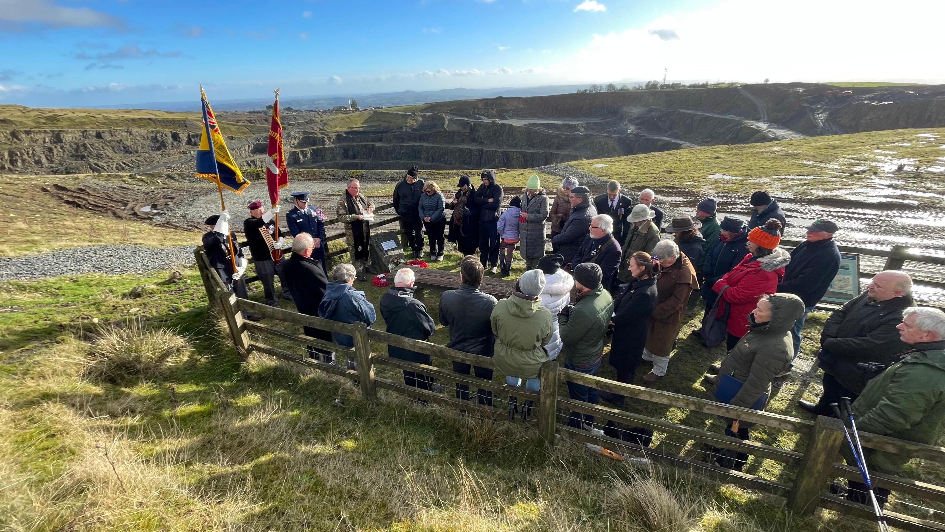 Dozens of people stand in a fenced off area around a memorial stone. There are servicemen with large flags, and in the background is a huge quarry and green hills.