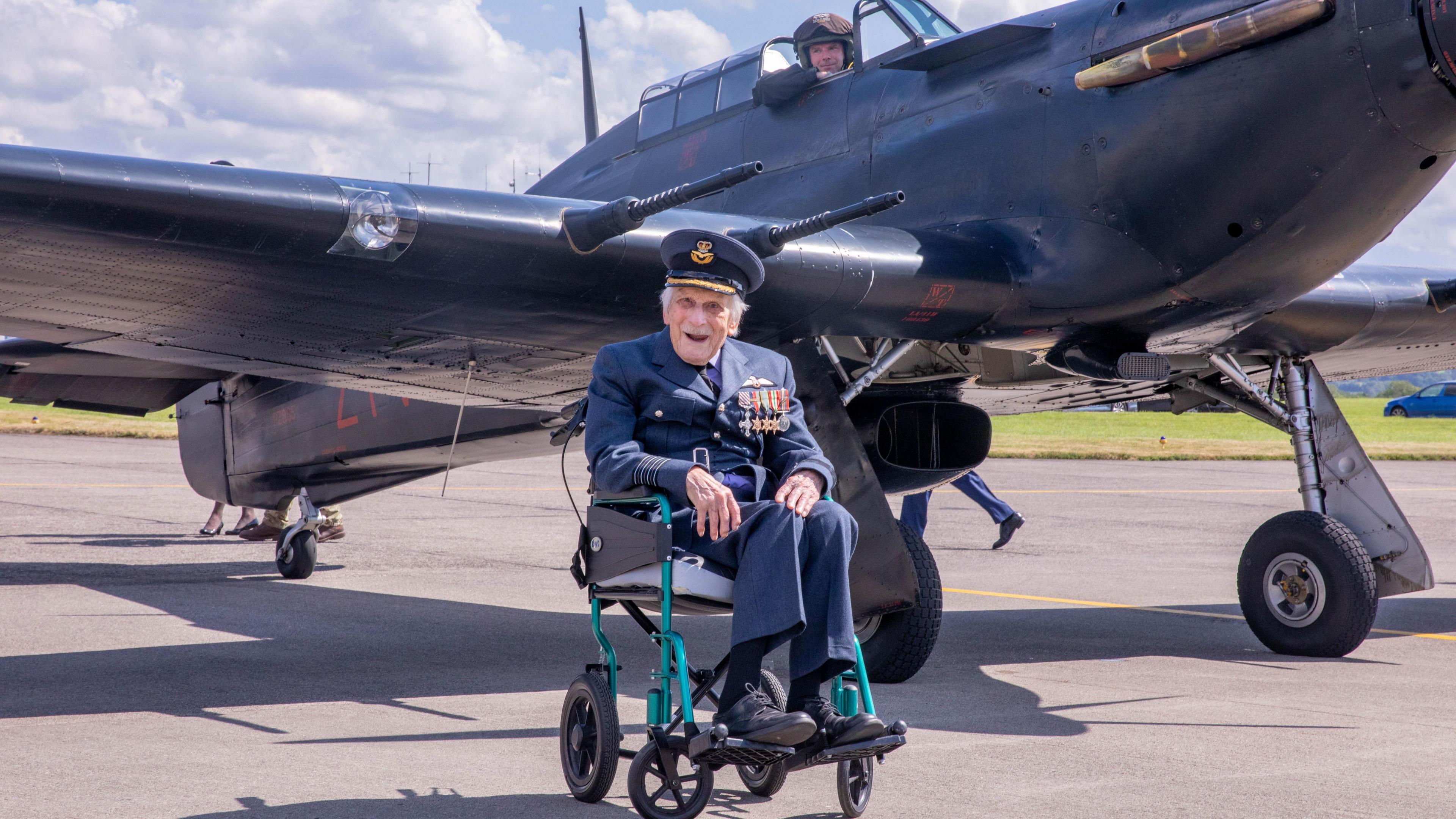 John 'Paddy' Hemingway sits in a wheelchair on an airstrip close to an RAF jet. He is smiling at the camera as he wears a full uniform