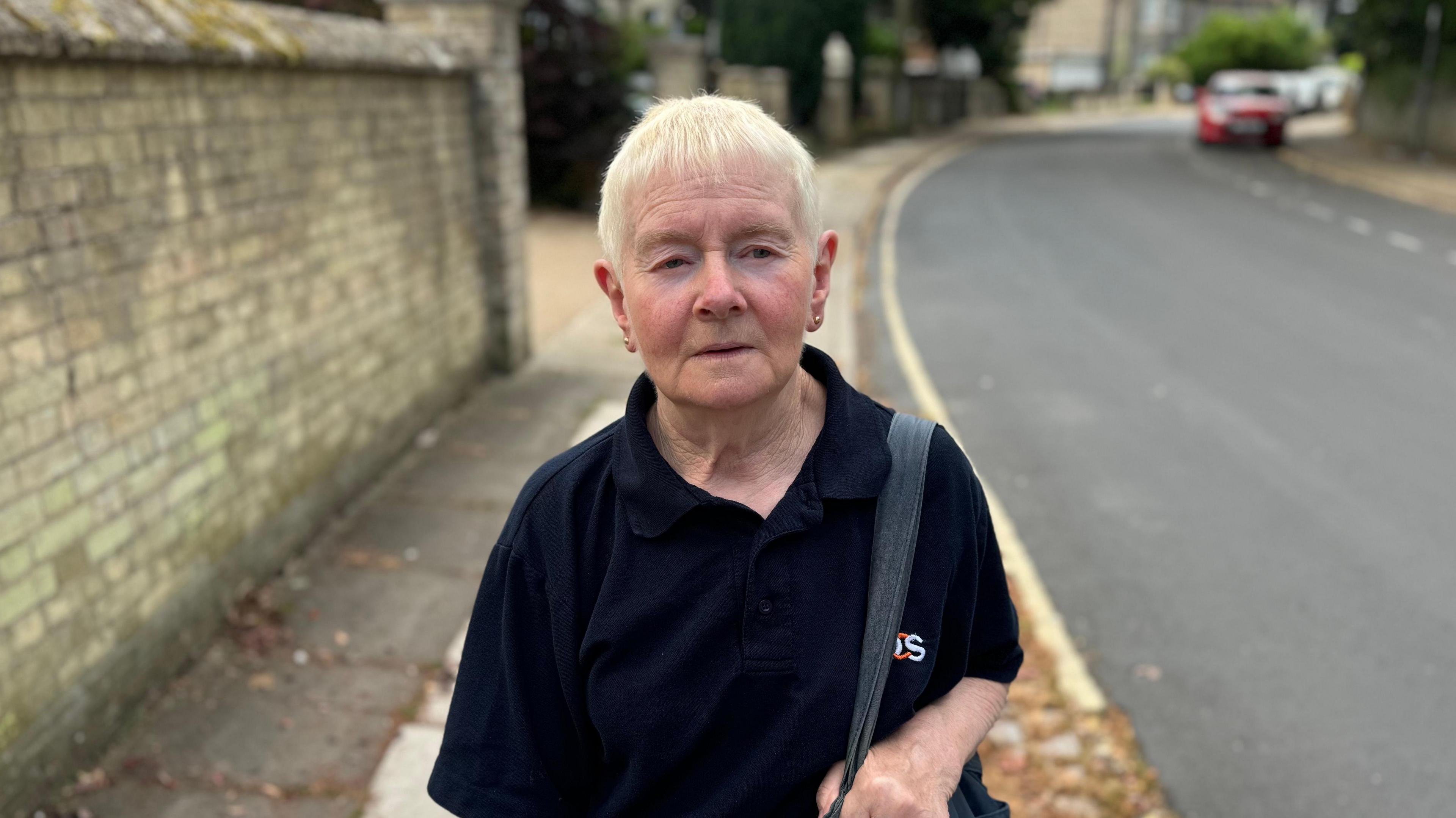 Mavis Cubit looking to the right of the camera, dressed in a dark polo neck shift and carrying a handbag. She has short blonde hair and is standing on the pavement in a street. 