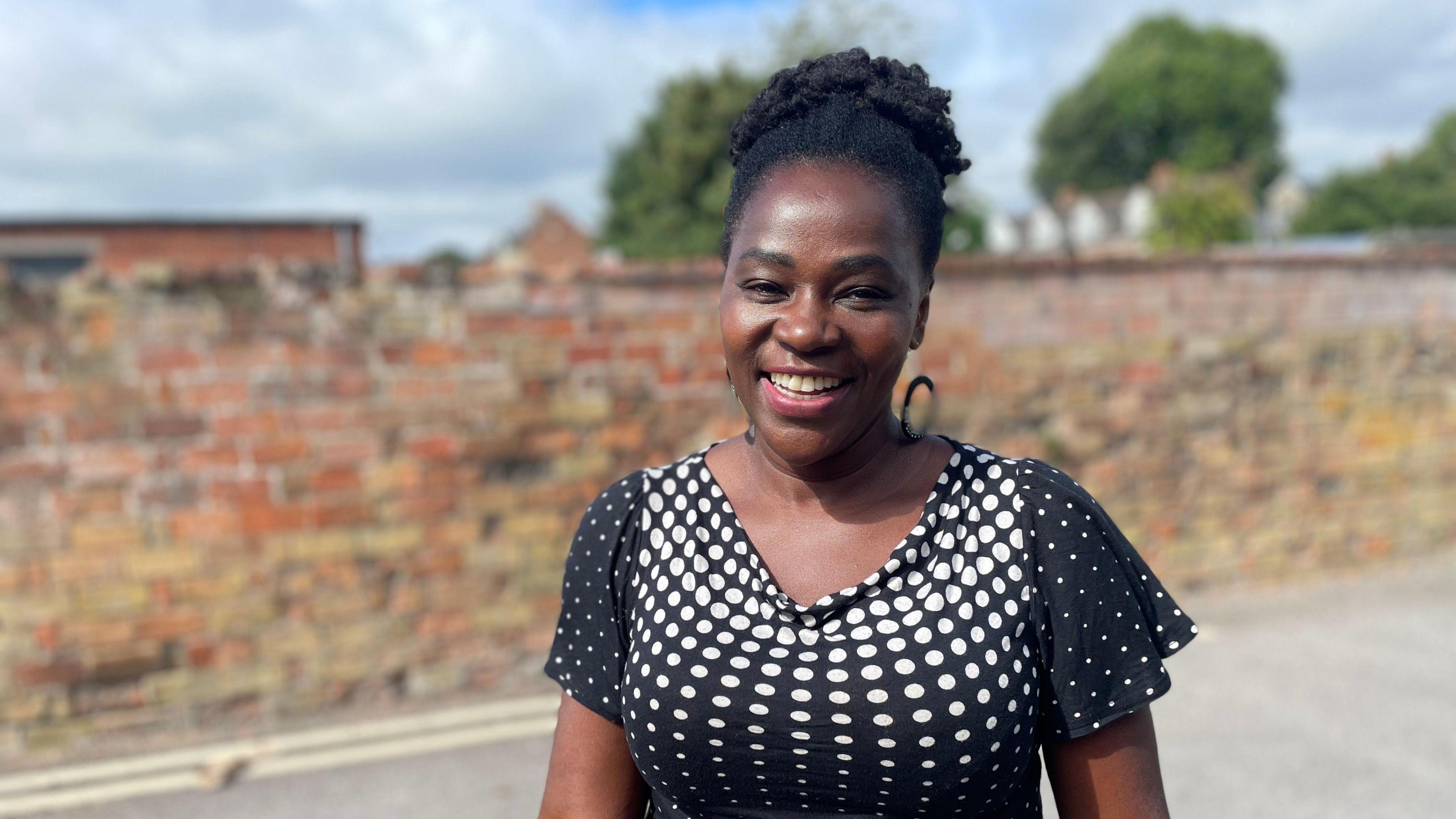 Angela Cook smiles at the camera. She is wearing a white and back spotty top and has her hair up. She is standing in front of a wall.