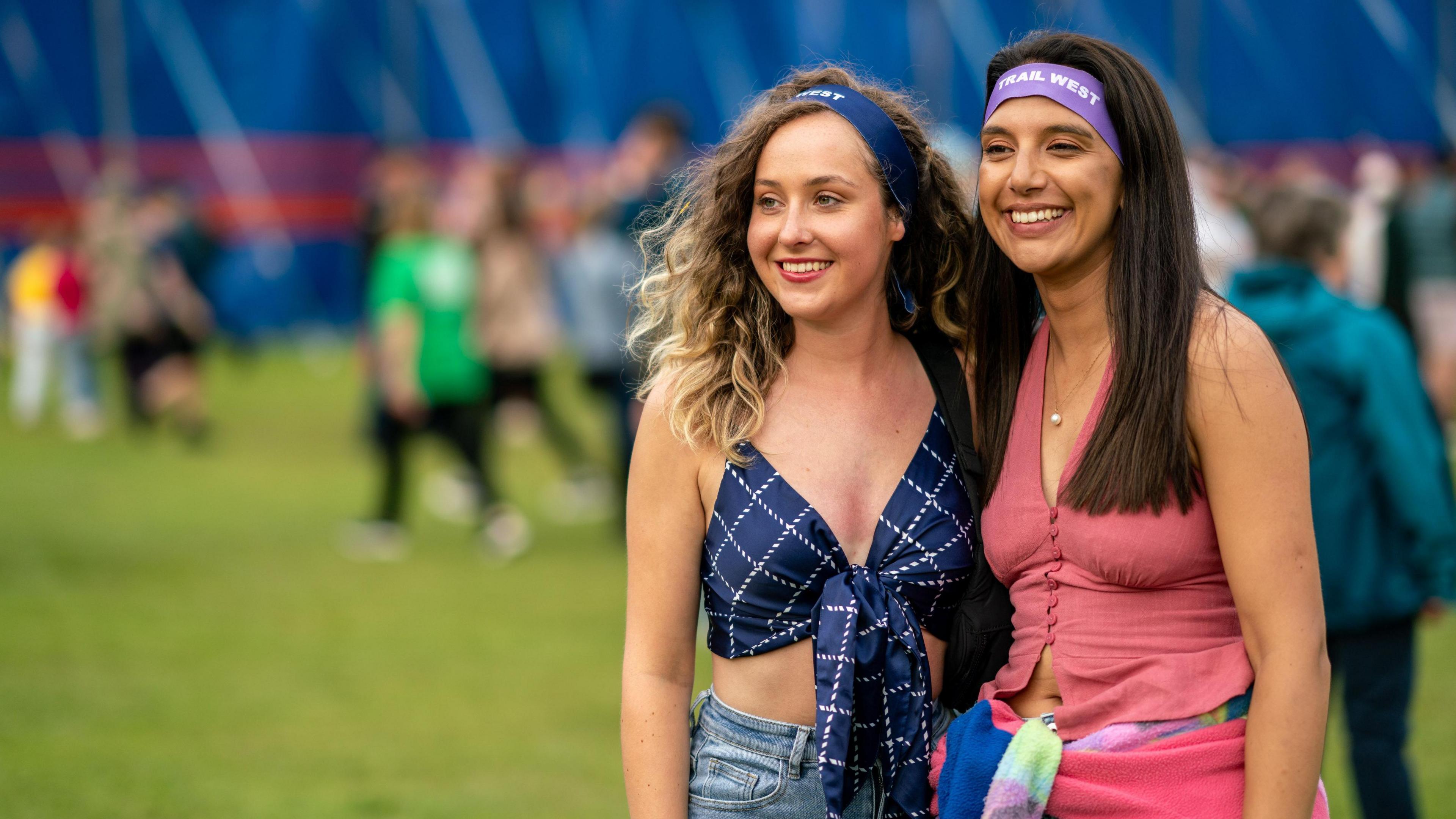 Two girls at the festival. One has long, curly blonde and brown hair and the other long, brown hair. They are both wearing crop tops and headbands