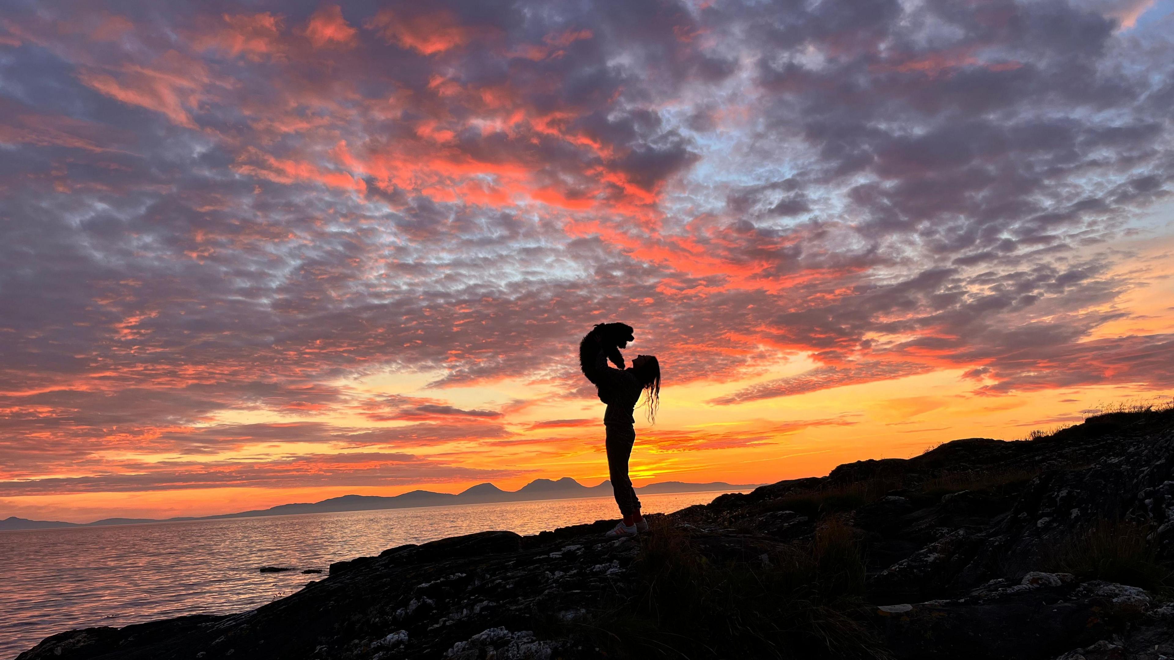 Vivid sky showing blue, orange, red and yellow at sunset with the shadowed outline of a woman holding a dog in the air, standing by the sea