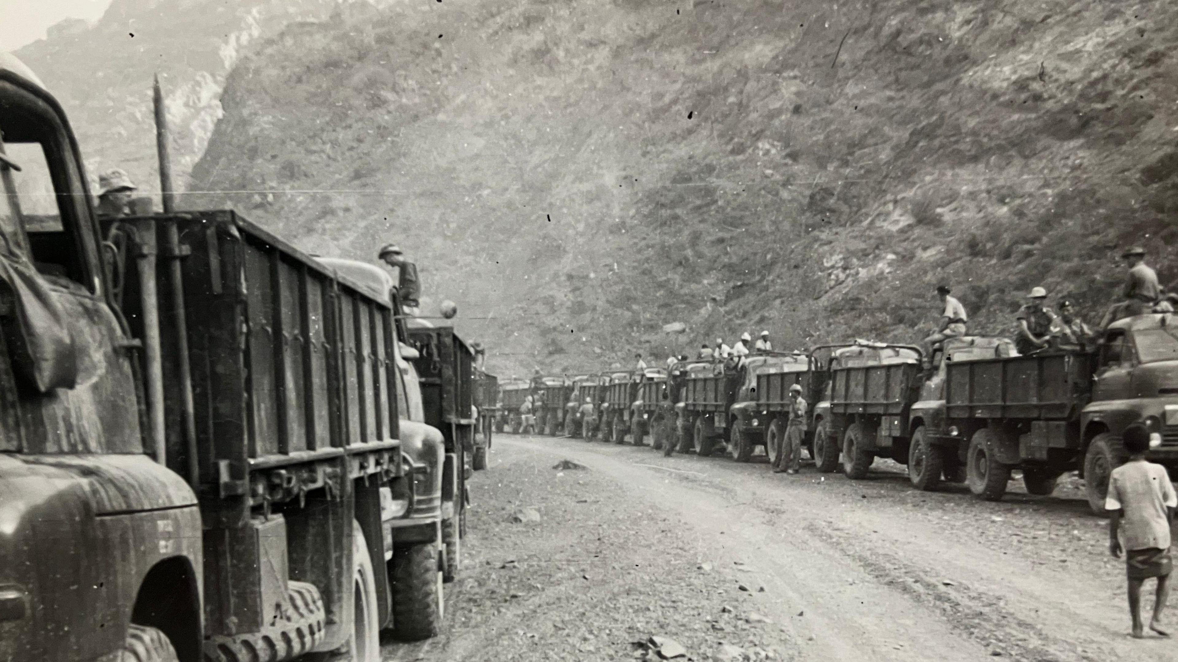 Black and white photo showing a military convoy of 15 or more trucks carrying troops and supplies. The road is rough and dusty and surrounded by a steep gorge.